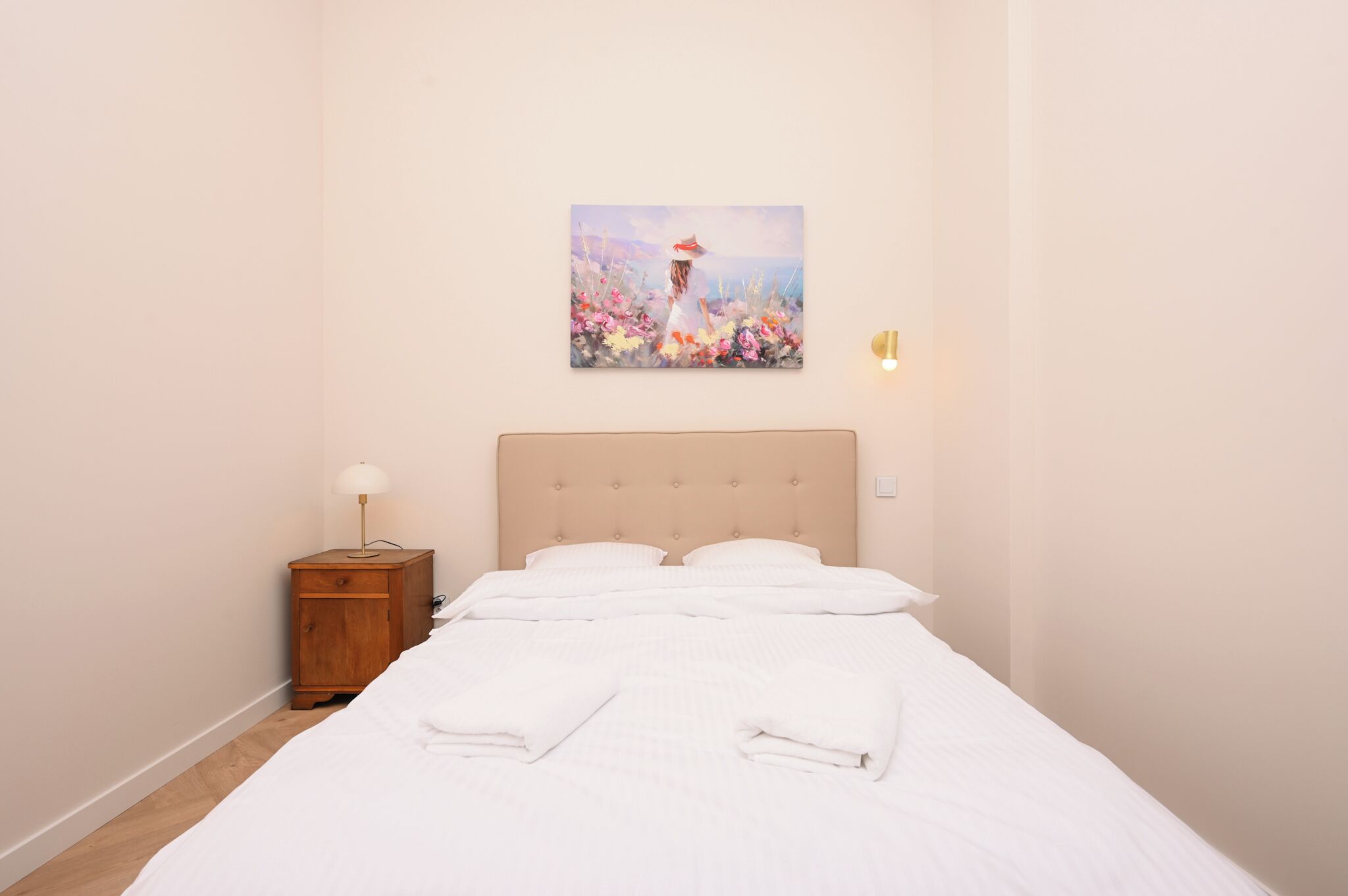 Ein ordentlich gemachtes Bett mit weißer Bettwäsche, ein Nachttisch aus Holz mit einer Lampe und ein Bild einer Frau auf einem Feld an der Wand.