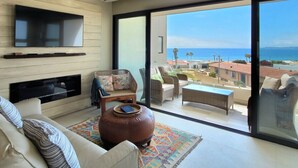 Una sala de estar moderna con un televisor grande, asientos cómodos, una chimenea y un balcón con vistas al mar.