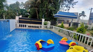A swimming pool with a waterfall feature, a white fence, and a blue-roofed building in the background. - Sami Resort  (Tirupattur)