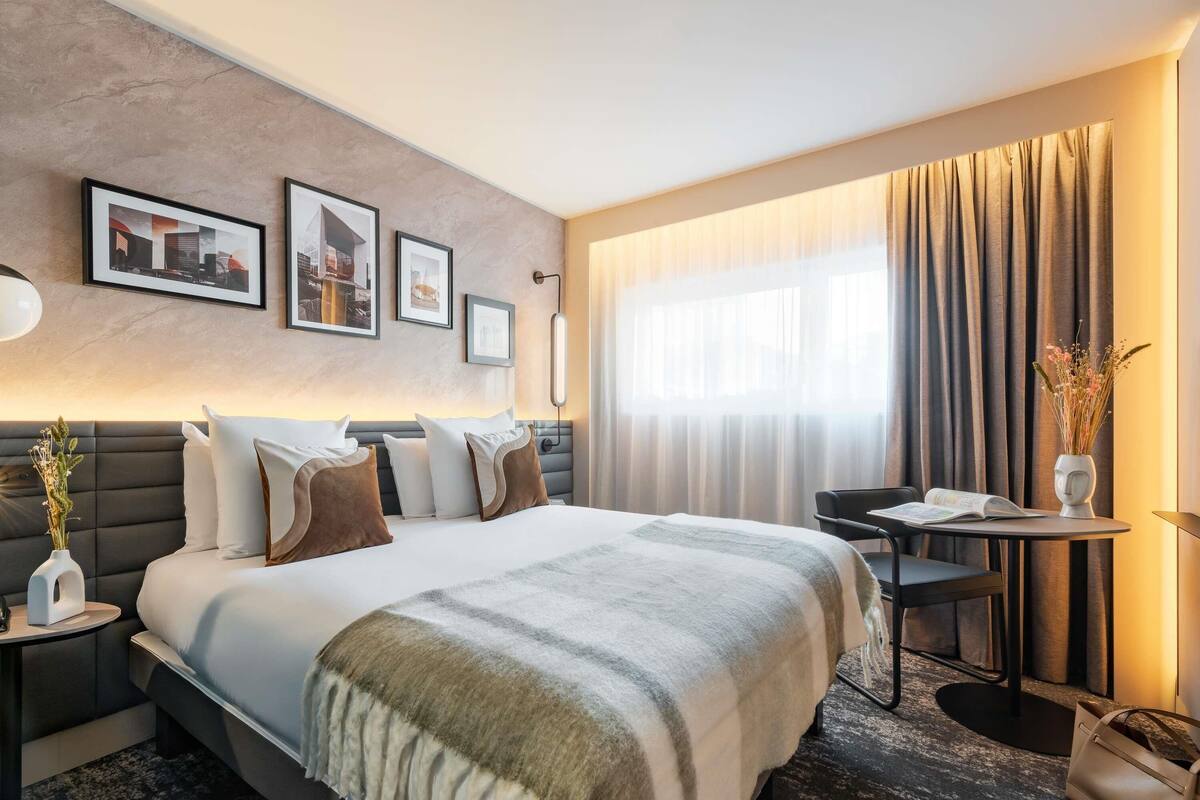 A hotel room with a large bed, a desk, a chair, a vase with flowers, and framed pictures on the wall.