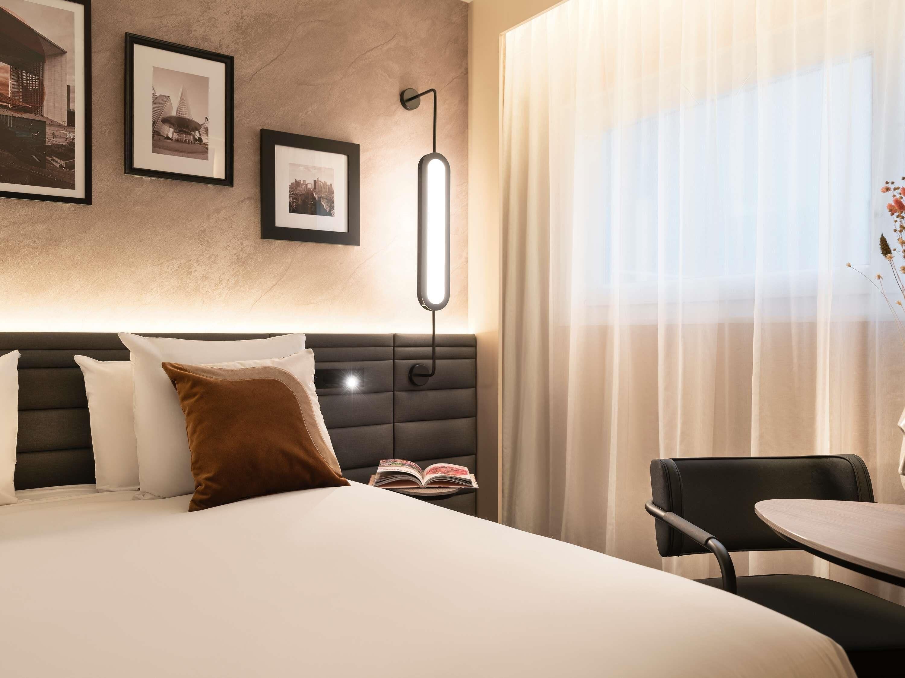A hotel room with a bed, a bedside table with a book, a chair, a lamp, and framed pictures on the wall.