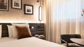 Une chambre d’hôtel avec un lit, une table de chevet sur laquelle se trouve un livre, une chaise, une lampe et des tableaux encadrés accrochés au mur.