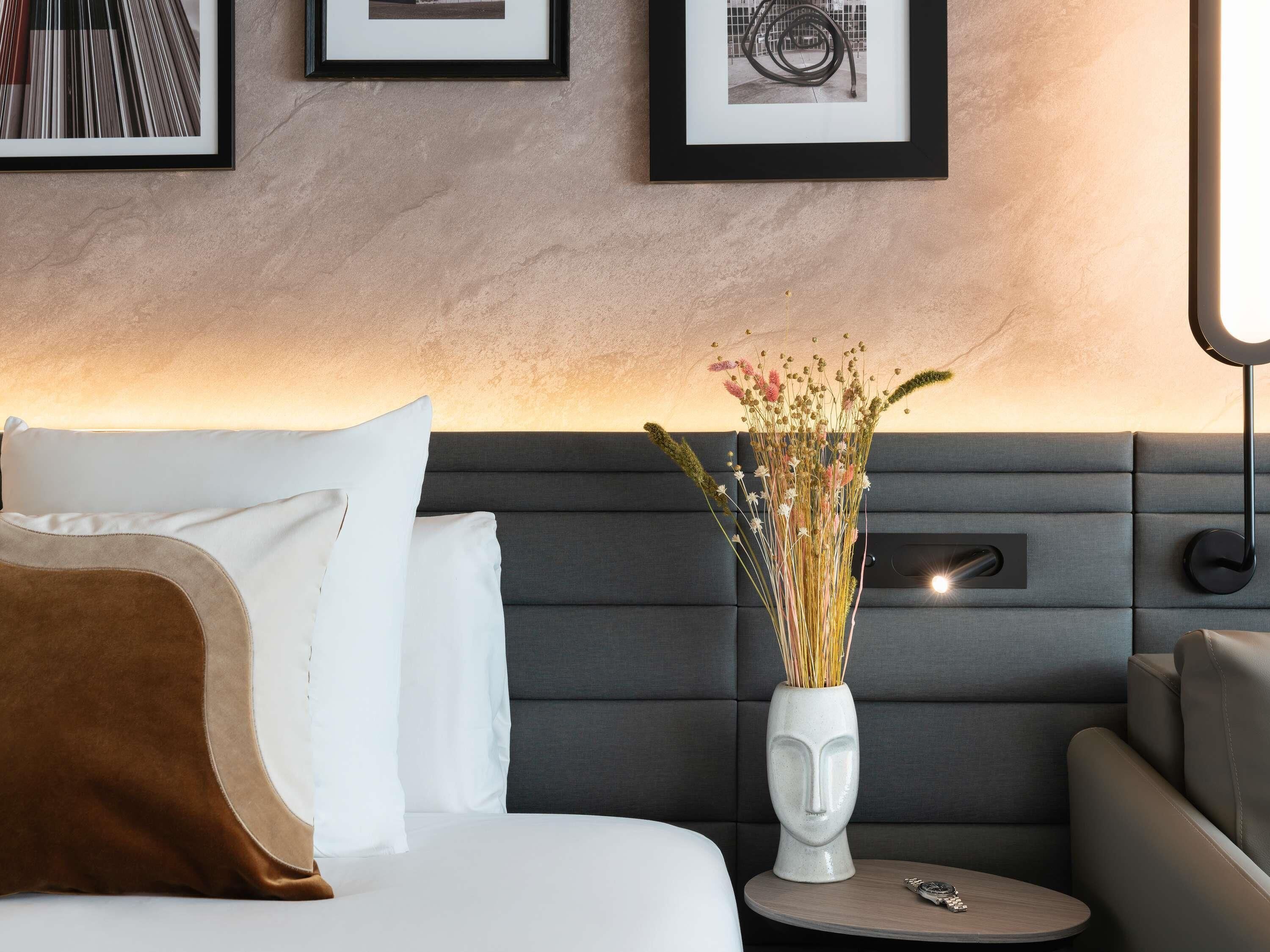 A modern hotel room with a bed, a sofa, a vase with dried flowers, and framed pictures on the wall.