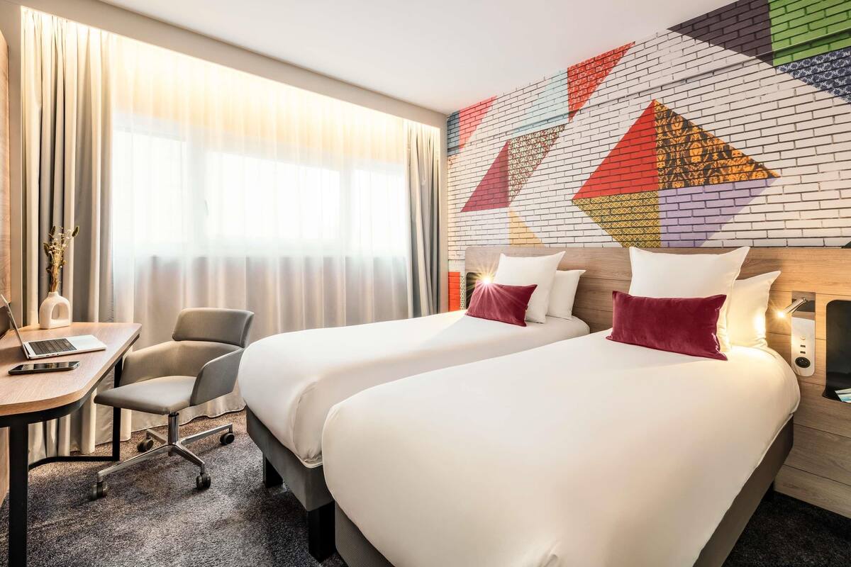A hotel room with two beds, a desk, a chair, and a large window with curtains.