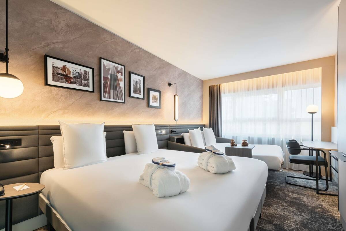 Ein Hotelzimmer mit einem großen Bett, Nachttischen, einem Schreibtisch und einem Stuhl. An der Wand hängen gerahmte Bilder und ein Fenster mit Vorhängen.