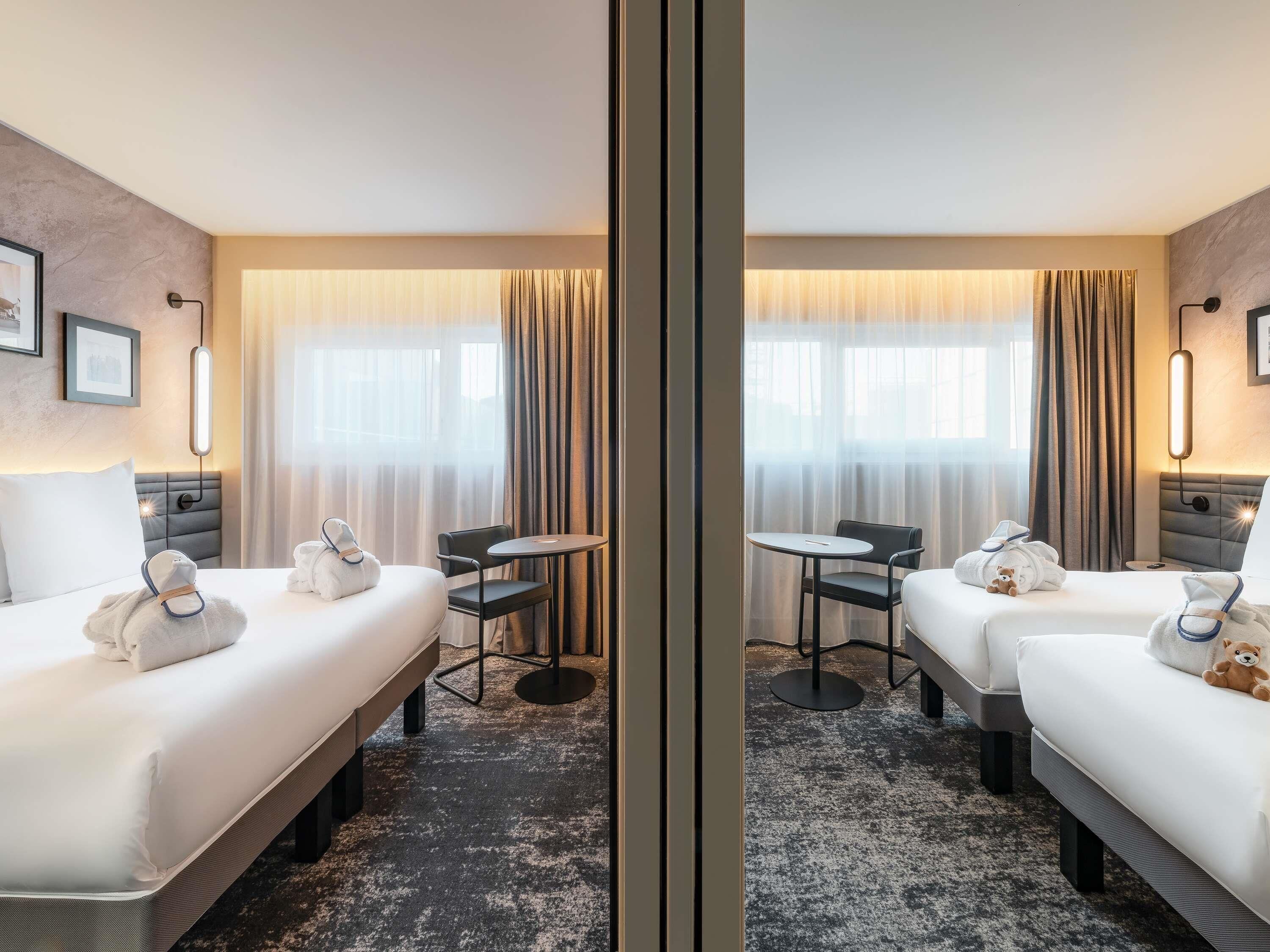 A hotel room with two beds, a small table, chairs, a sofa, and a large window with curtains.