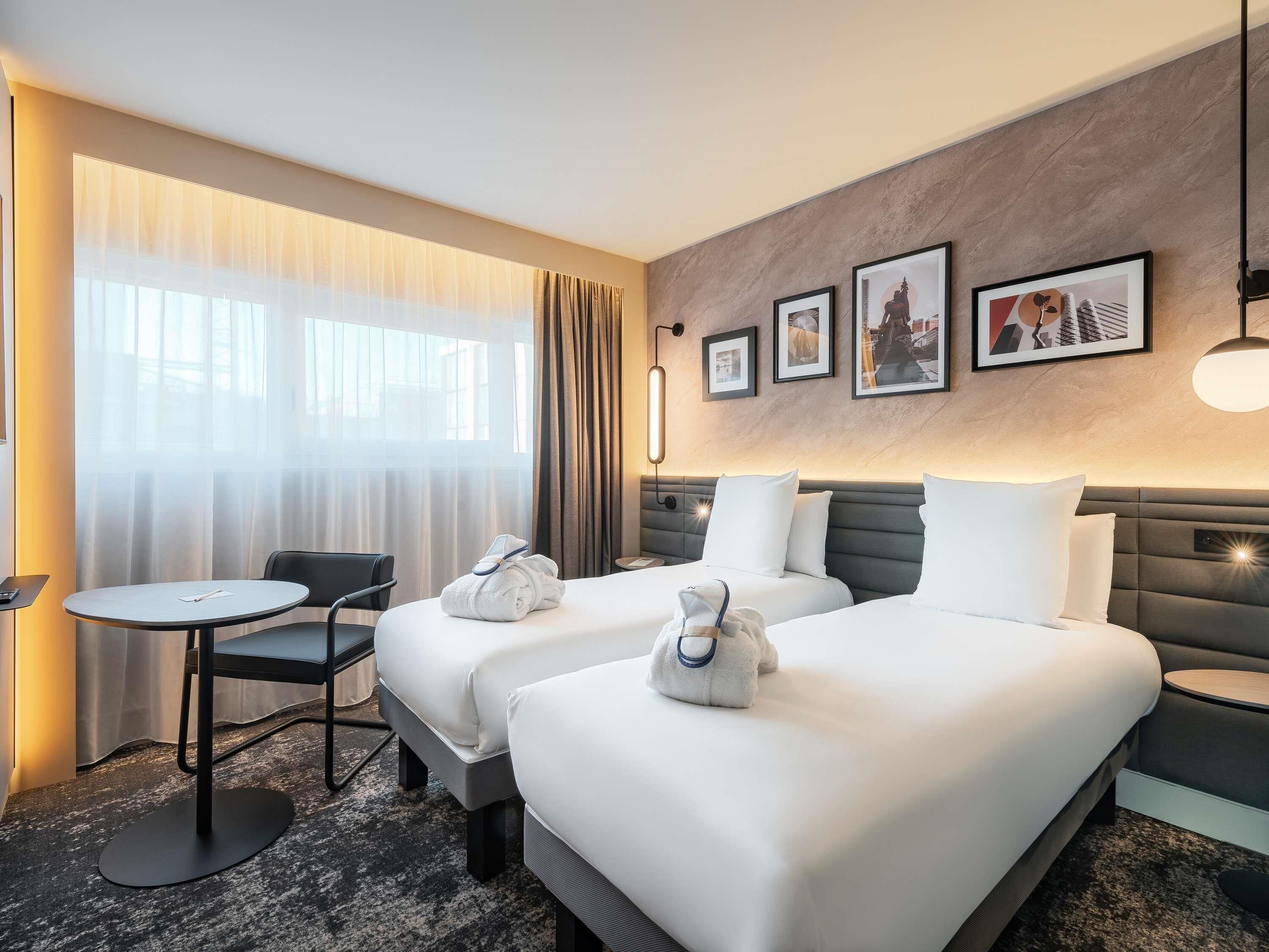 A hotel room with two beds, a small table, a chair, and a large window with curtains.