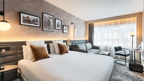 Une chambre d’hôtel moderne avec un grand lit, un canapé, un bureau et une chaise. Des tableaux encadrés ornent le mur et une lampe se trouve sur le bureau.