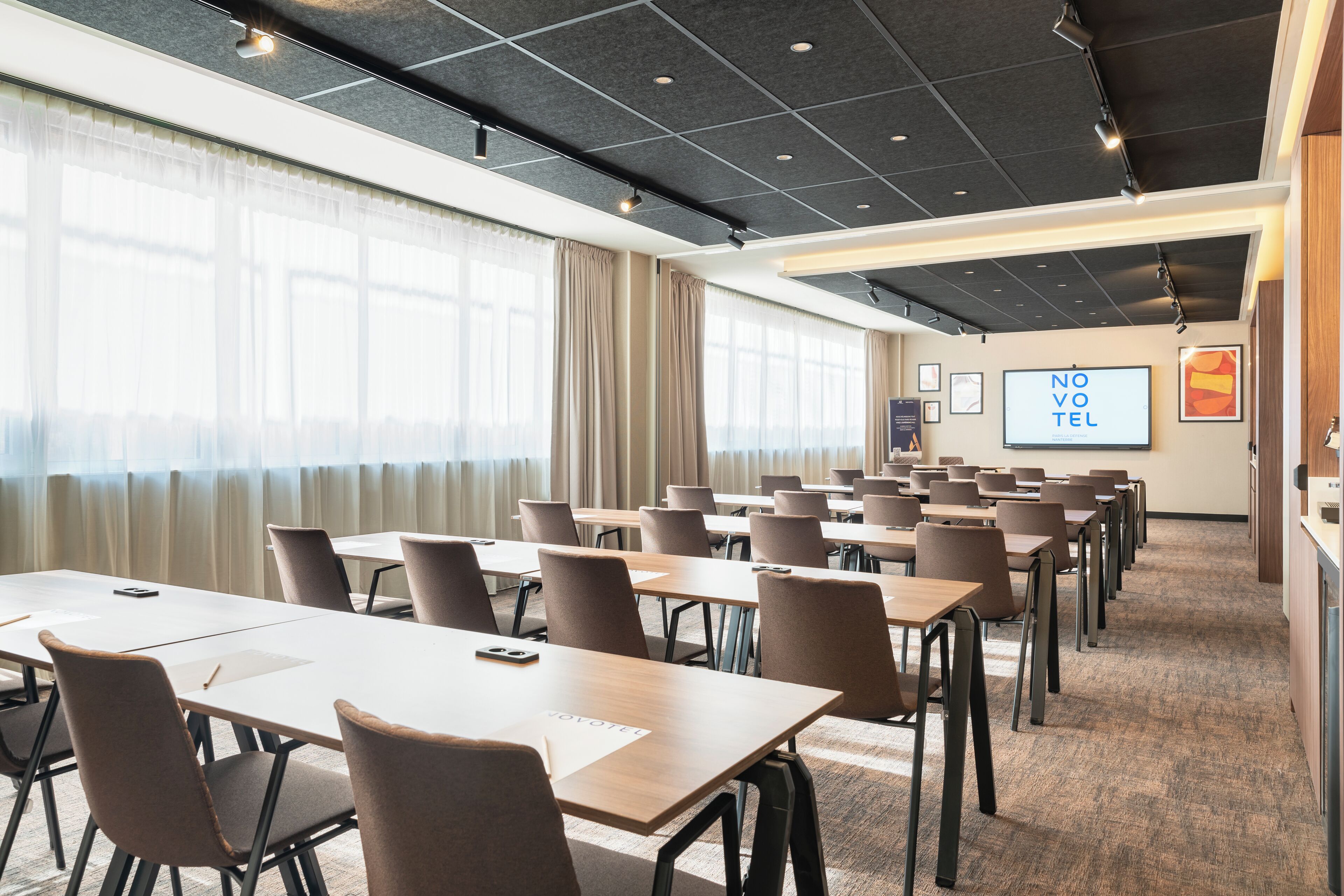 A conference room with multiple tables and chairs, large windows with curtains, and a ceiling with recessed lighting.