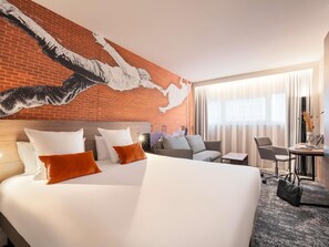 Habitación de hotel con una cama grande, un sofá, un escritorio y vistas a la ciudad a través del ventanal.