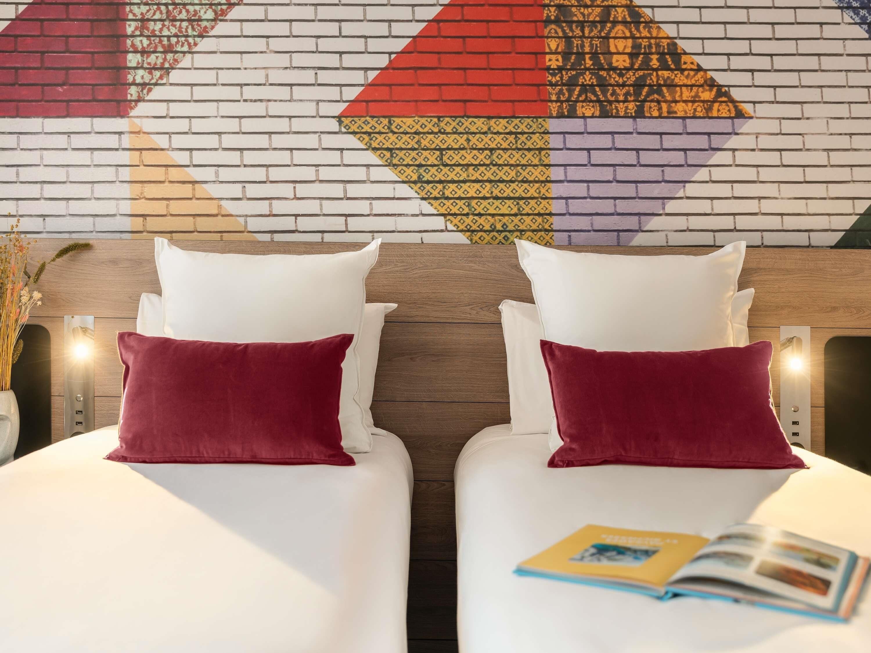 Two beds with white bedding and red pillows, a brick-patterned wall, and a wooden headboard.
