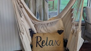 Een hangstoel met een kussen waarop de tekst 'Relax' staat, voor een raam met gordijnen en uitzicht op een tuin.