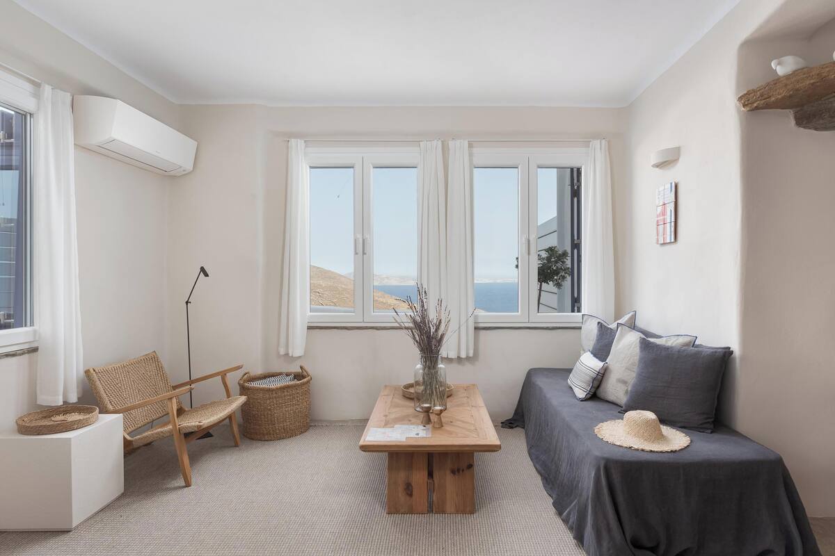 Quarto com vista para o mar, uma mesa de madeira com um vaso, uma cadeira de vime, um sofá com almofadas e um aparelho de ar condicionado branco.