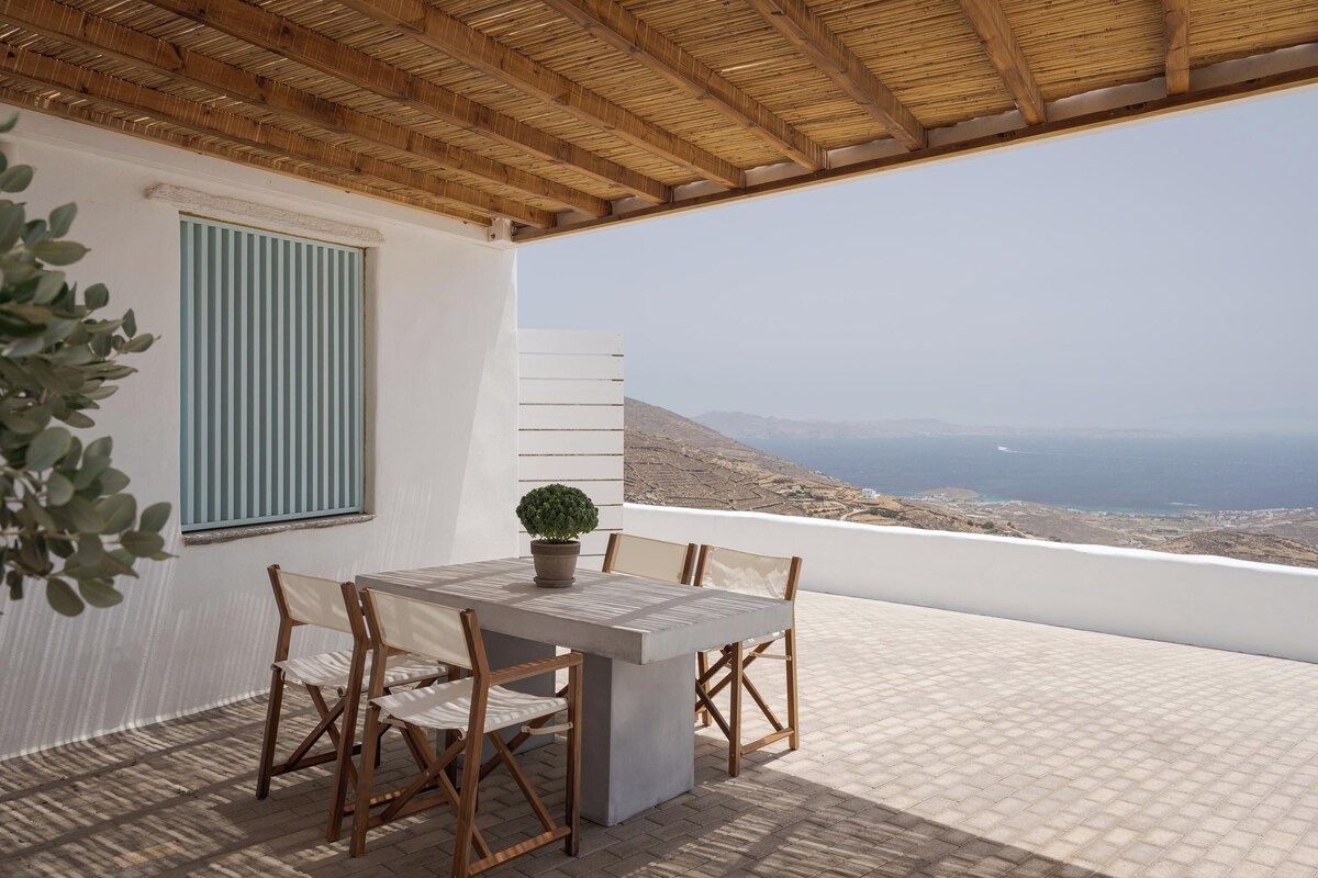 Una terraza con mesa y sillas, una planta en maceta y vistas al mar y las colinas.