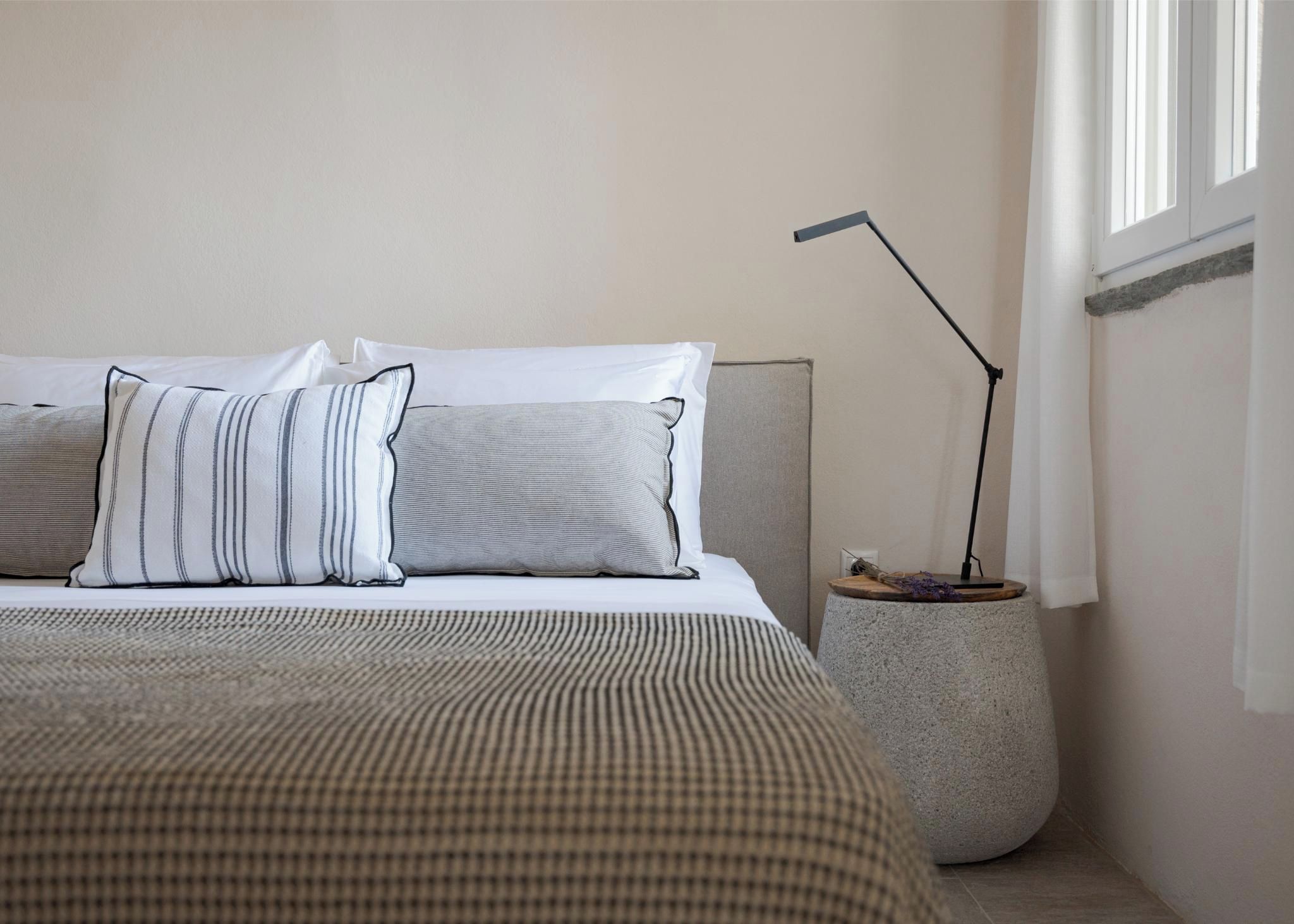 A neatly made bed with striped and solid pillows, a modern floor lamp, and a small side table with a vase of flowers.
