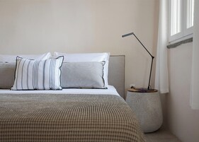 Ένα στρωμένο κρεβάτι με προσεγμένο τρόπο, με ριγέ και μονόχρωμα μαξιλάρια, ένα μοντέρνο φωτιστικό δαπέδου και ένα μικρό βοηθητικό τραπεζάκι με ένα βάζο λουλούδια.