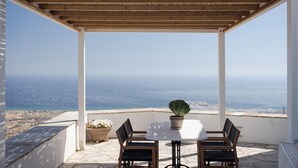 En overdækket terrasse med bord og stole, en potteplante og udsigt til kystlinjen.