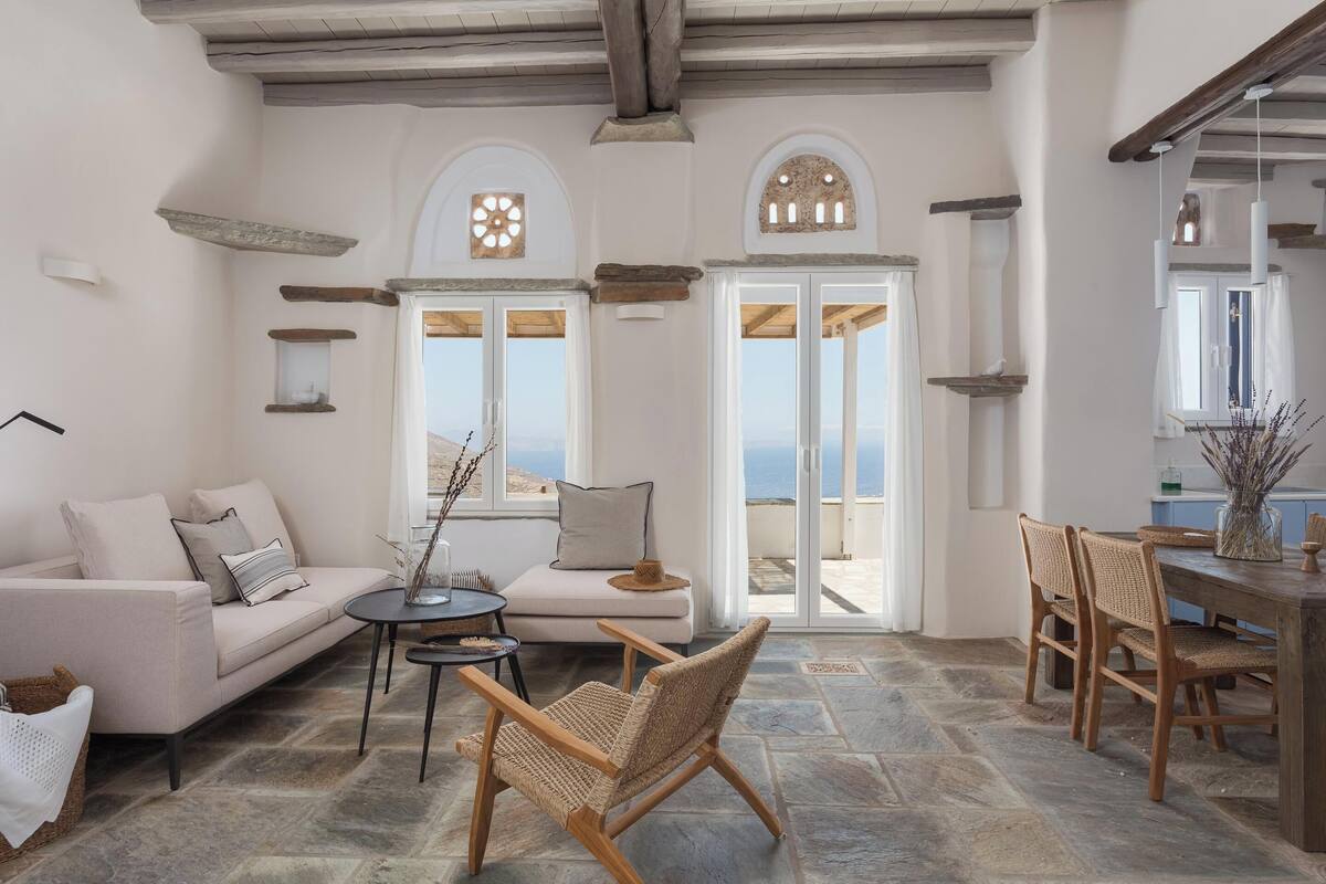Uma sala de estar ampla com um sofá, uma poltrona de vime, uma mesa de jantar com cadeiras e vista para o mar através de grandes portas.