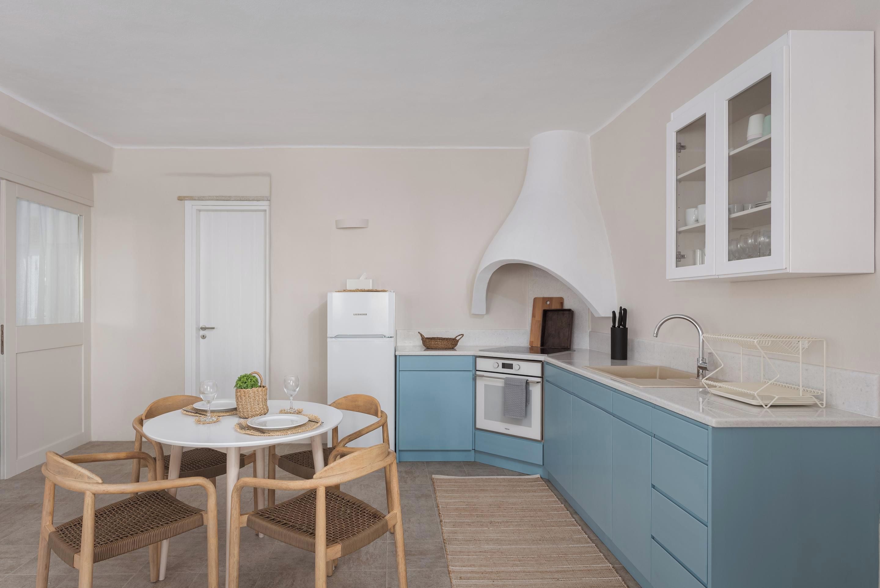 Une cuisine compacte avec un réfrigérateur blanc, un four encastré, un évier et un meuble bleu. On y trouve une table à manger avec des chaises et un petit tapis sur le sol.