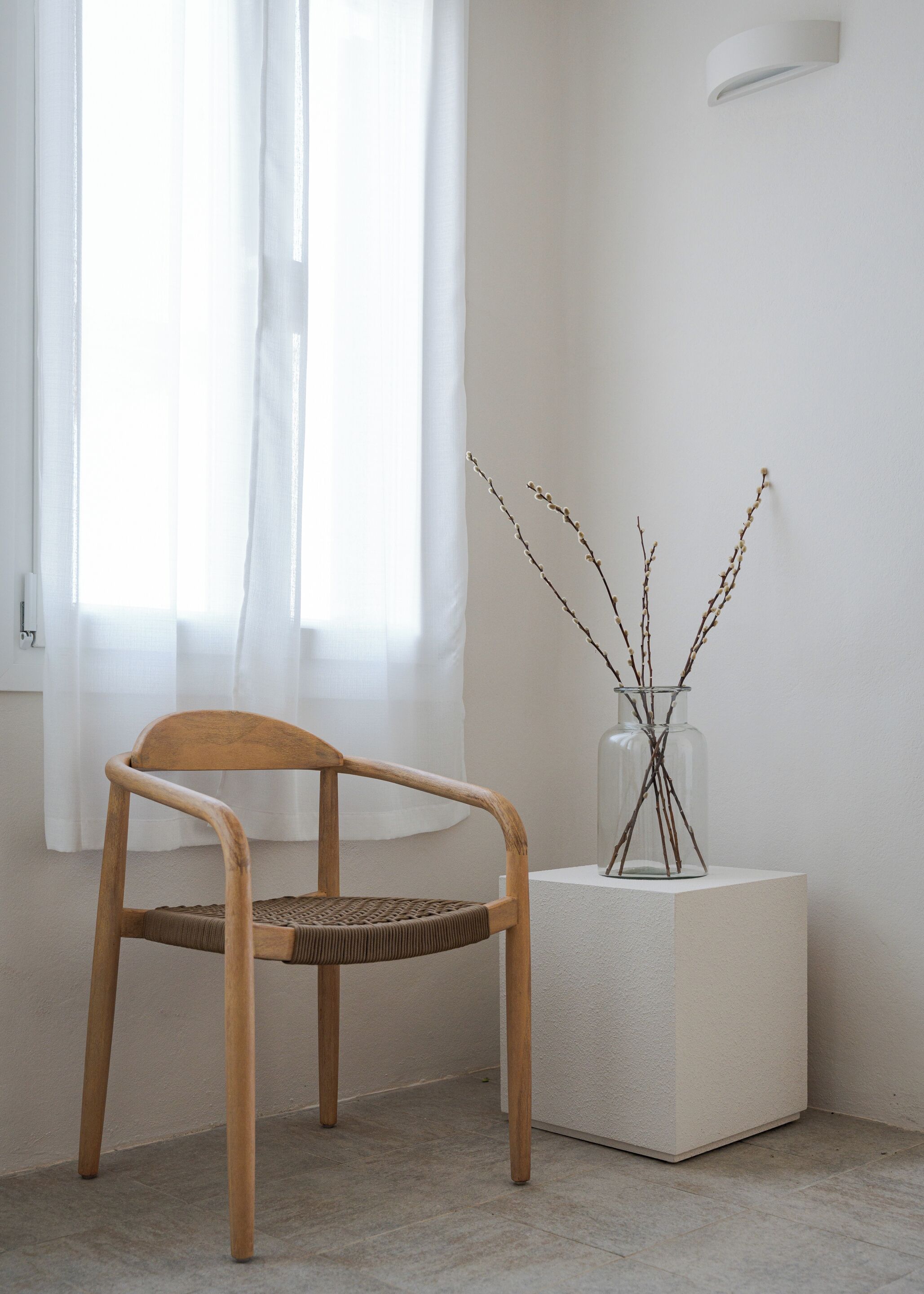 Uma cadeira de madeira com assento e encosto trançados, posicionada em frente a uma janela com cortinas brancas leves. Um vaso de vidro com galhos está colocado sobre um pedestal branco ao lado da cadeira.