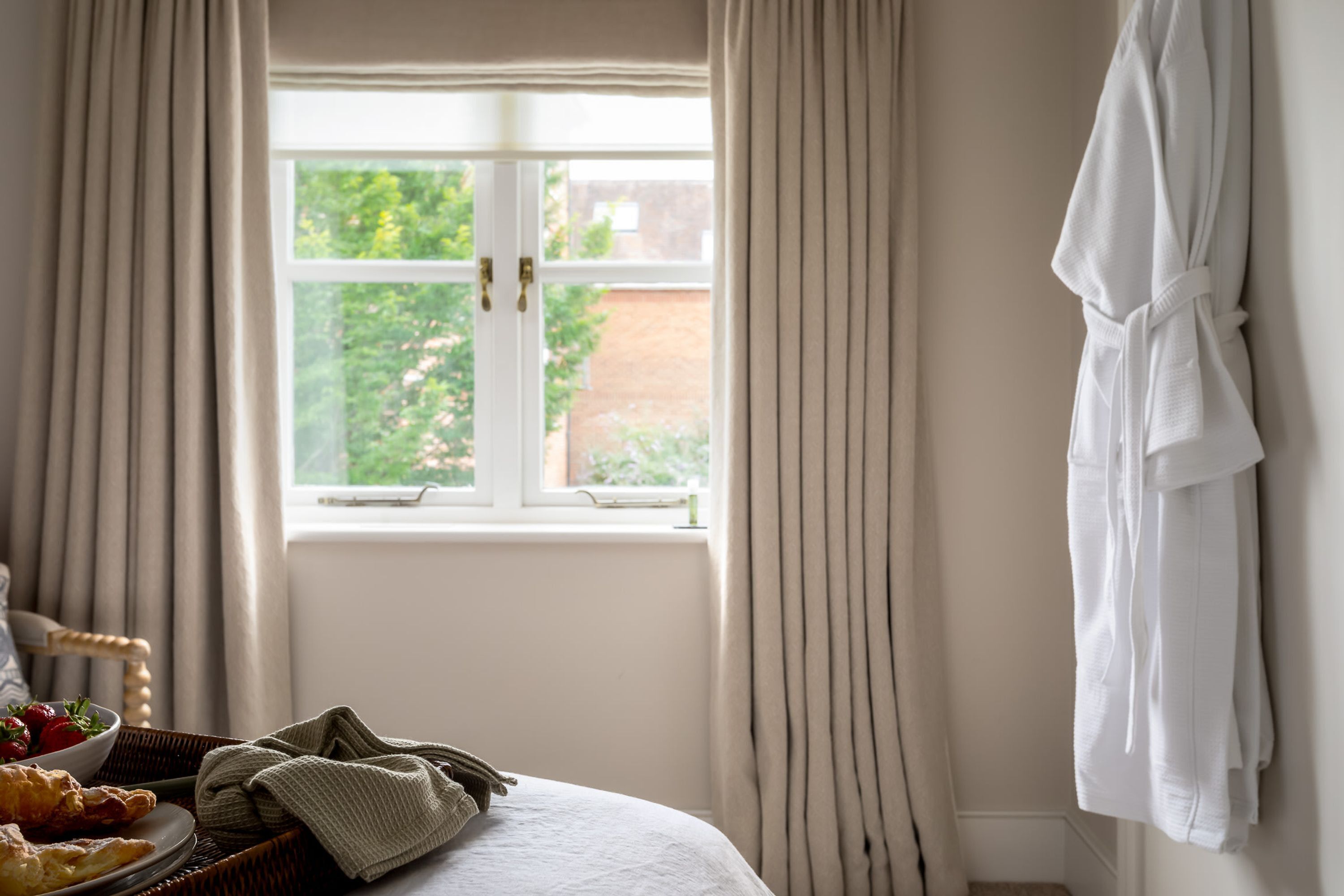 Una habitación de hotel con una cama, una ventana con cortinas, una bata colgada en la pared y una bandeja de desayuno sobre la cama.