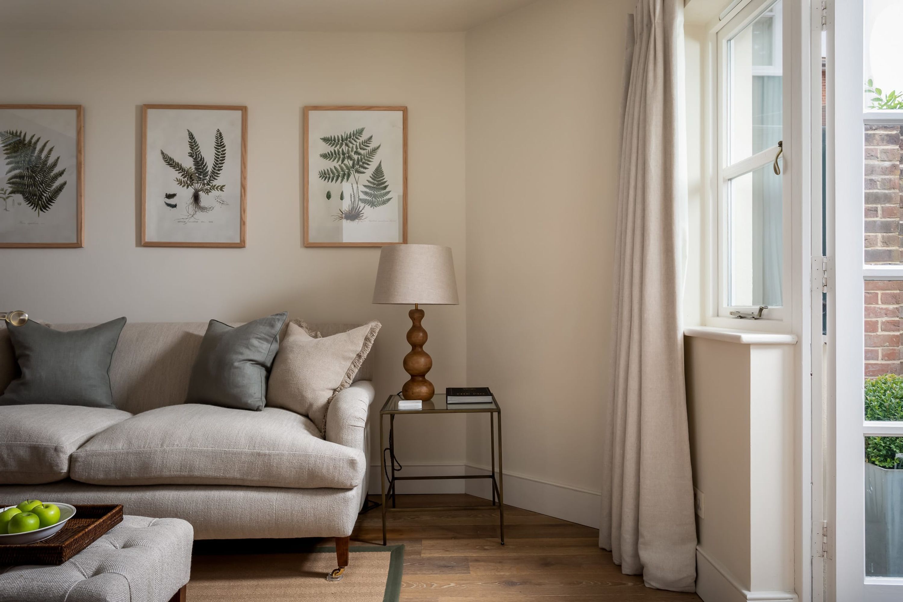 Un salon avec un canapé, une lampe, des reproductions botaniques encadrées accrochées au mur et une fenêtre avec des rideaux.