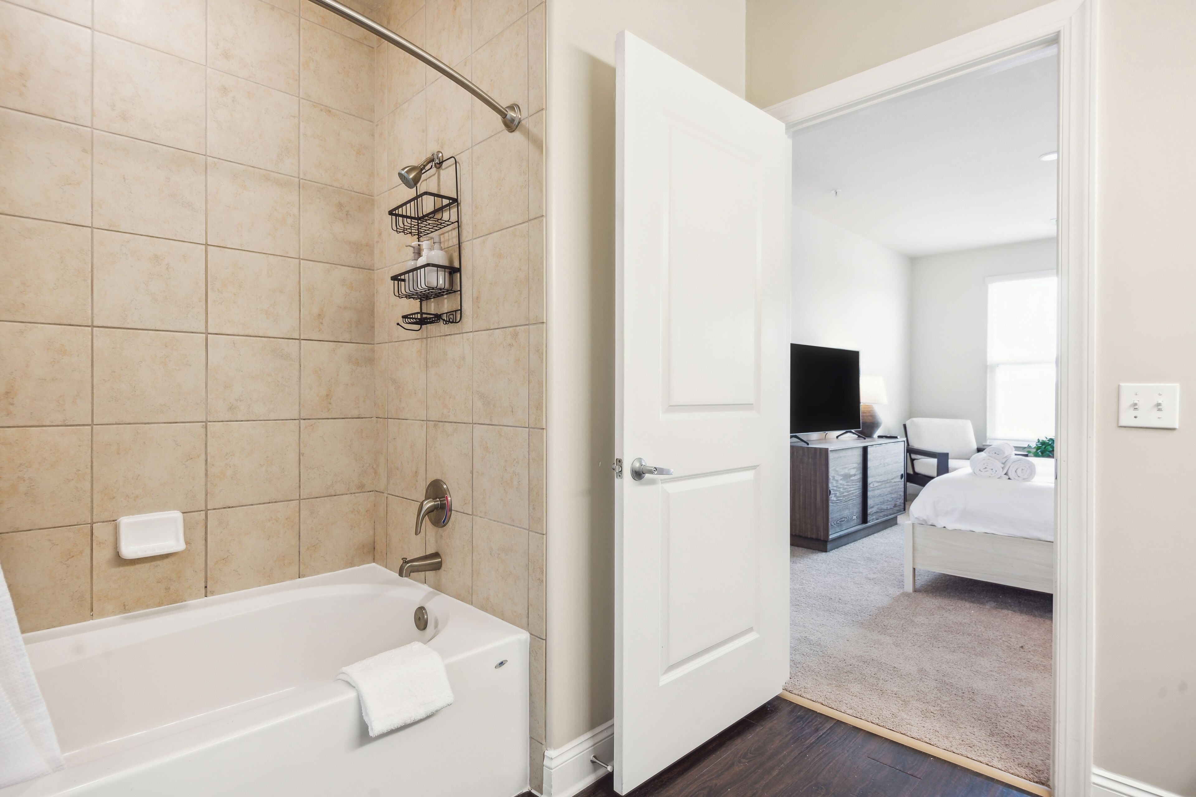 A modern bathroom with a bathtub, a shower, and a toilet. There is a white door leading to a bedroom with a bed and a TV.