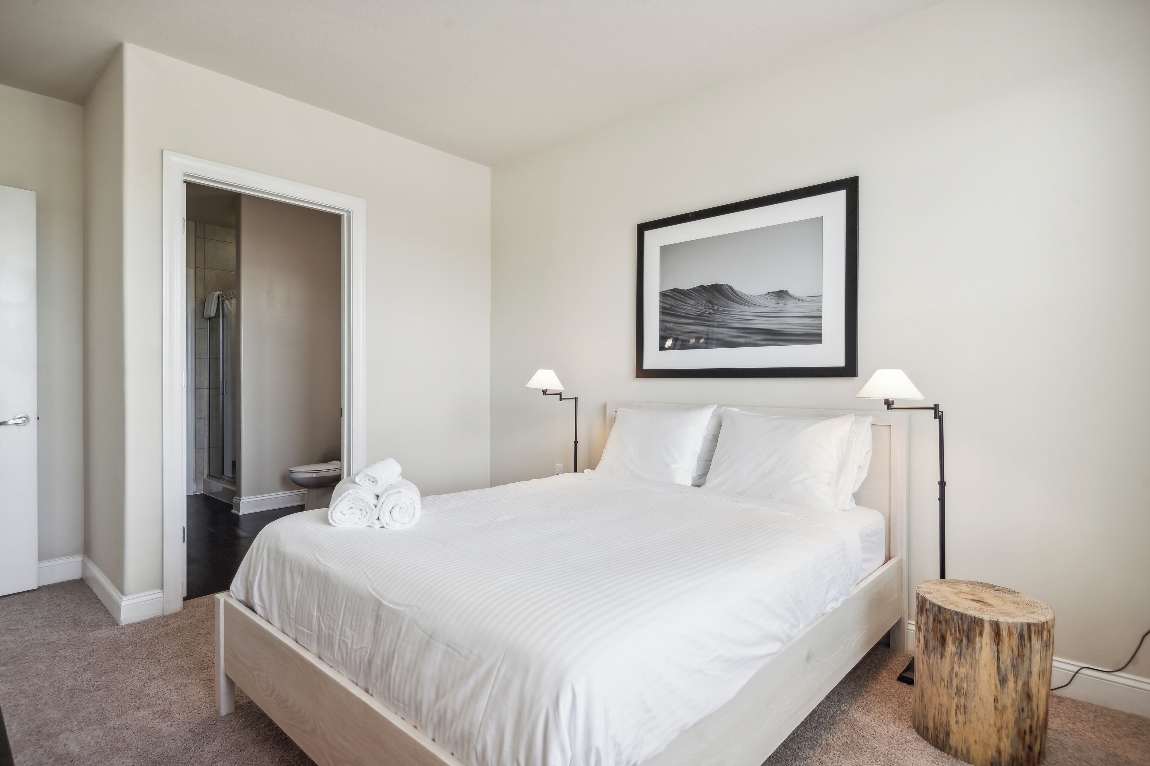 Quarto com uma cama, mesa de cabeceira, abajur, quadro na parede e porta que dá para um banheiro.