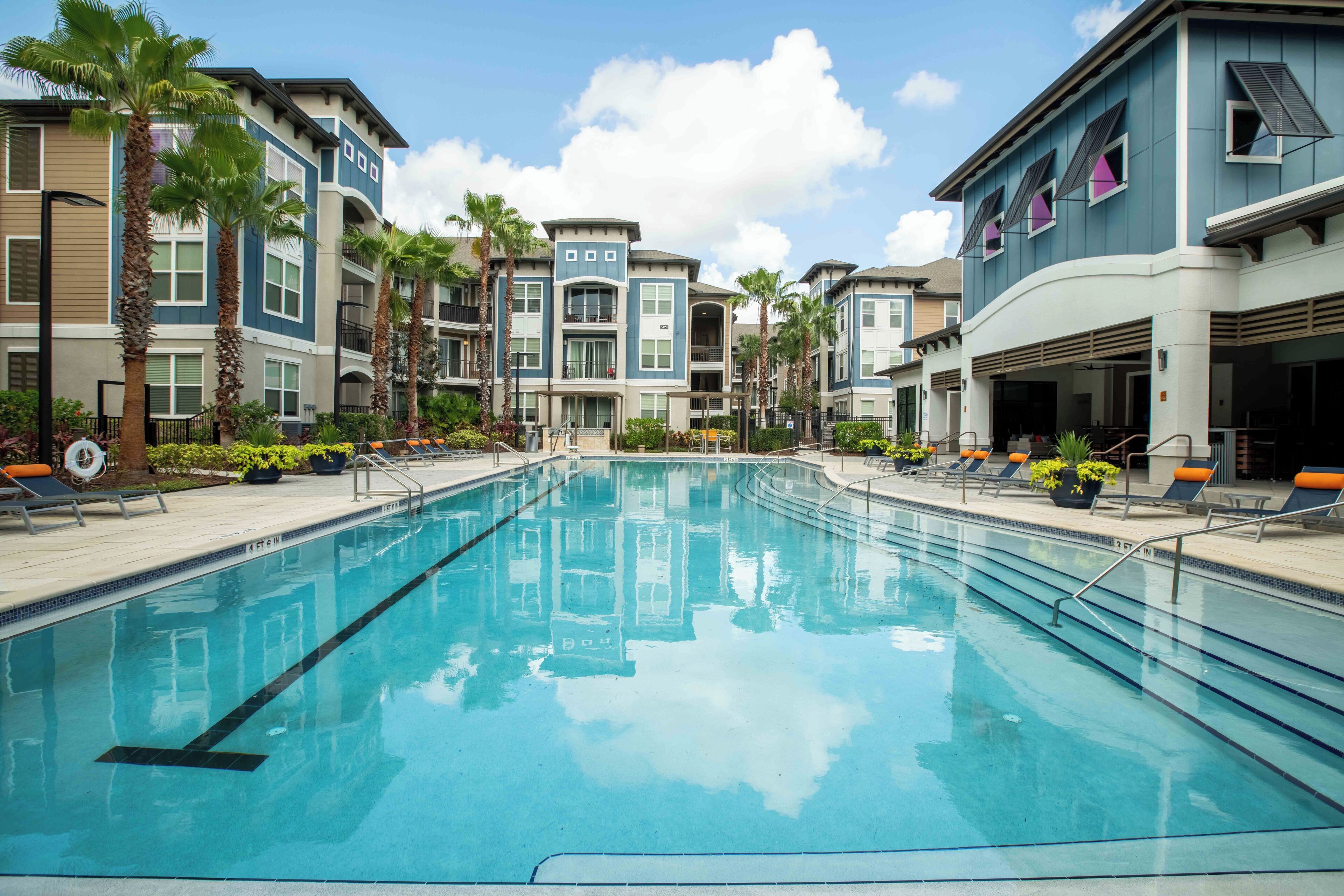 A swimming pool surrounded by multi-story buildings with balconies, palm trees, and lounge chairs.