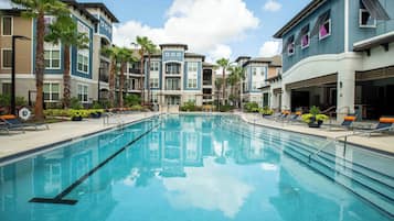 A swimming pool surrounded by multi-story buildings with balconies, palm trees, and lounge chairs.