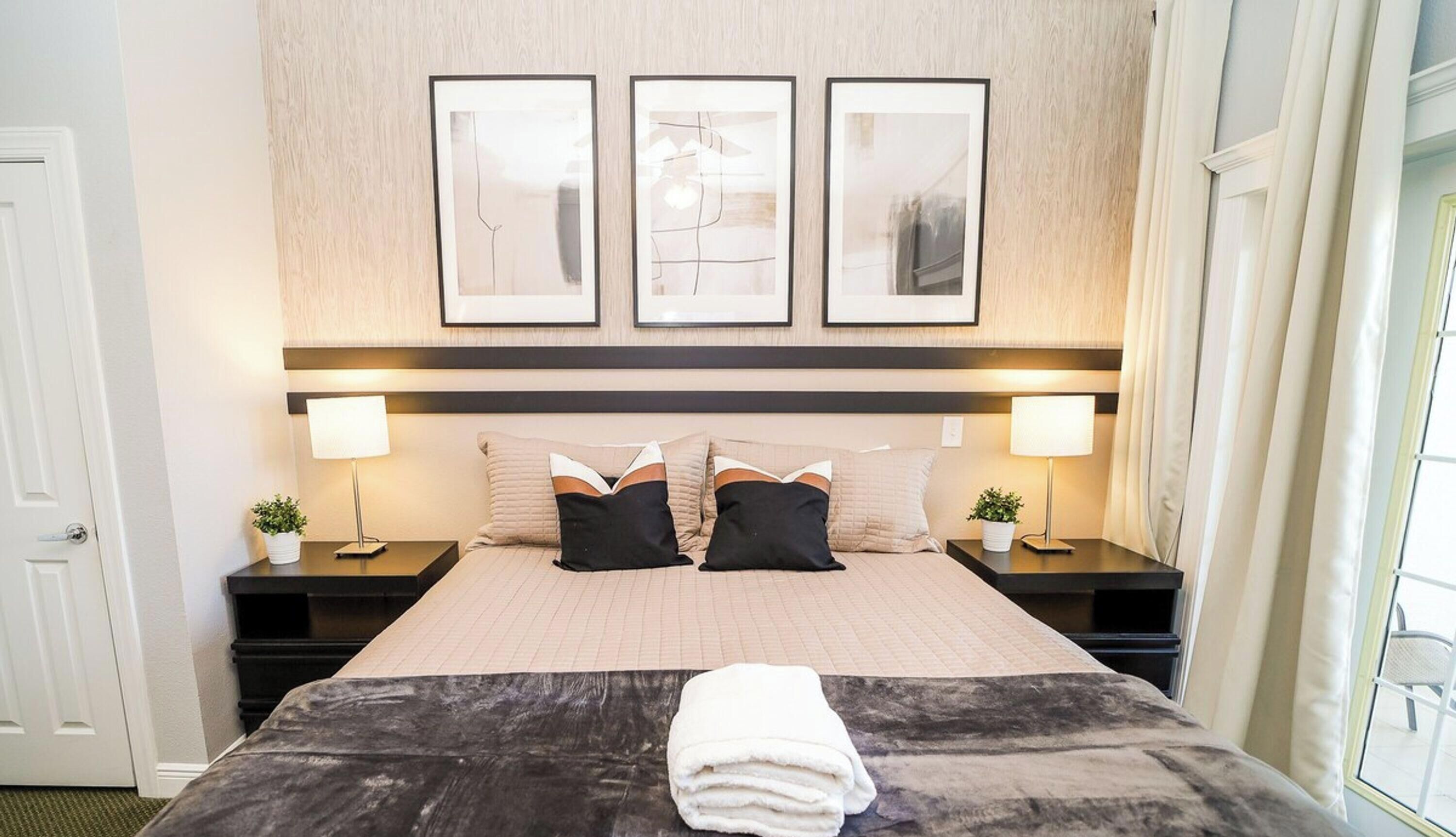 Une chambre d’hôtel comprenant un lit, deux tables de chevet, deux lampes, une fenêtre avec des rideaux et trois tableaux encadrés accrochés au mur.