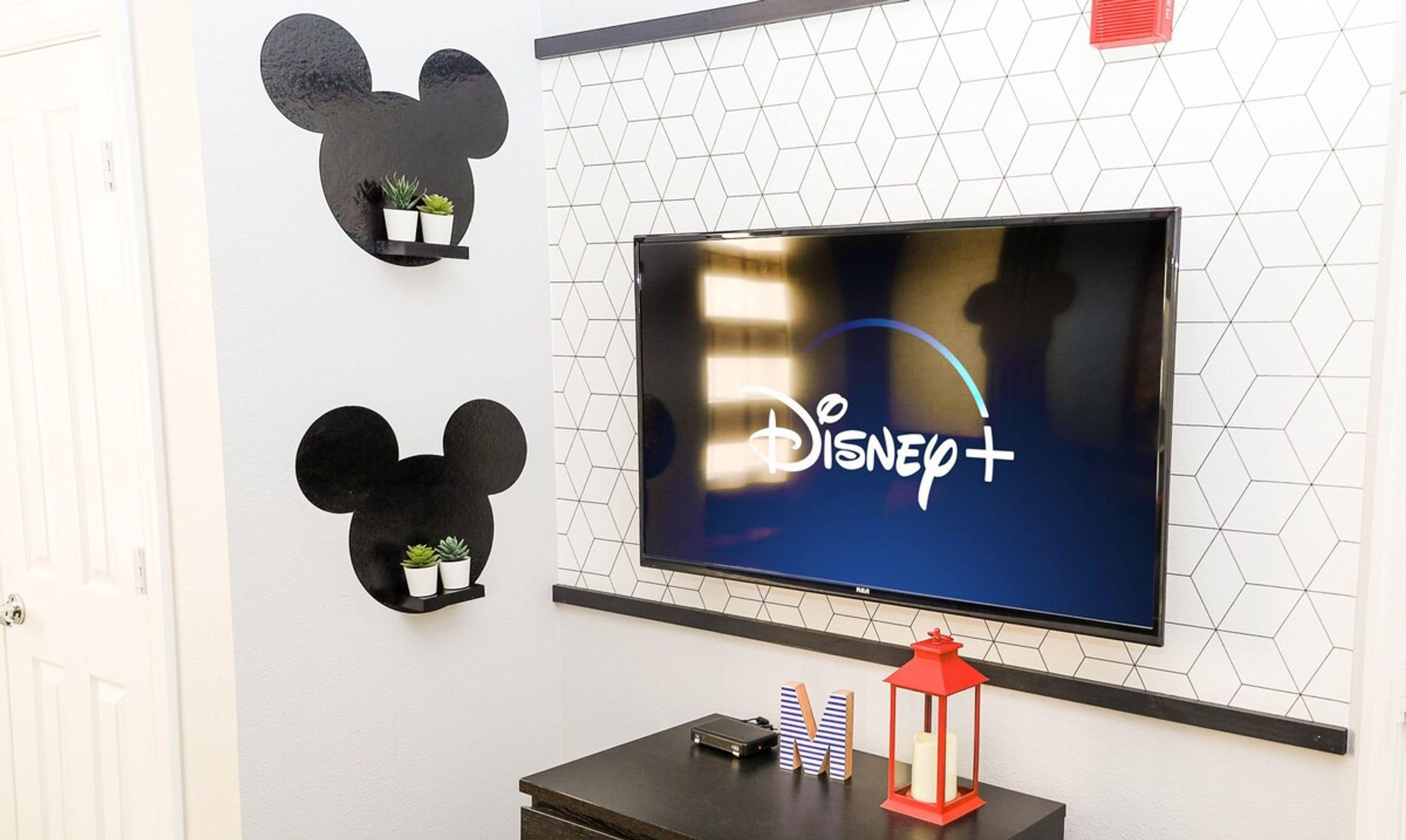 A television mounted on a wall displaying the Disney+ logo, with two black Mickey Mouse head decorations above it.