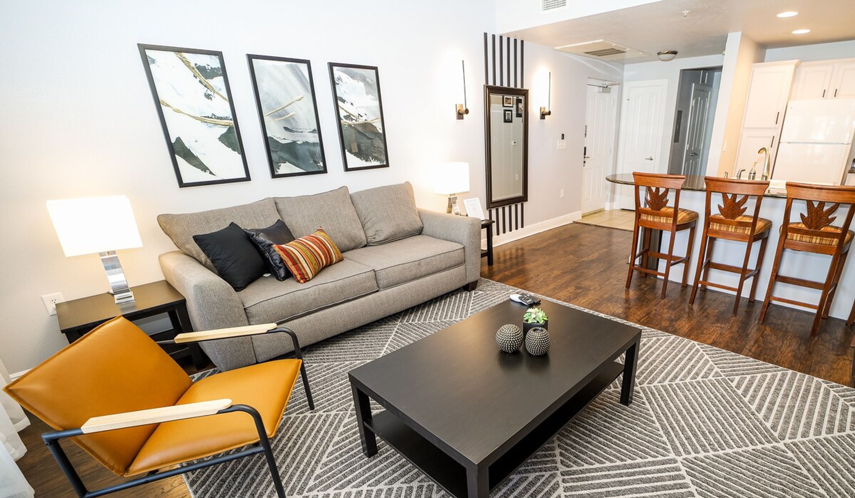 A modern living room with a grey sofa, a coffee table, and a patterned rug. There are three framed pictures on the wall, a lamp, and a dining area with wooden chairs.