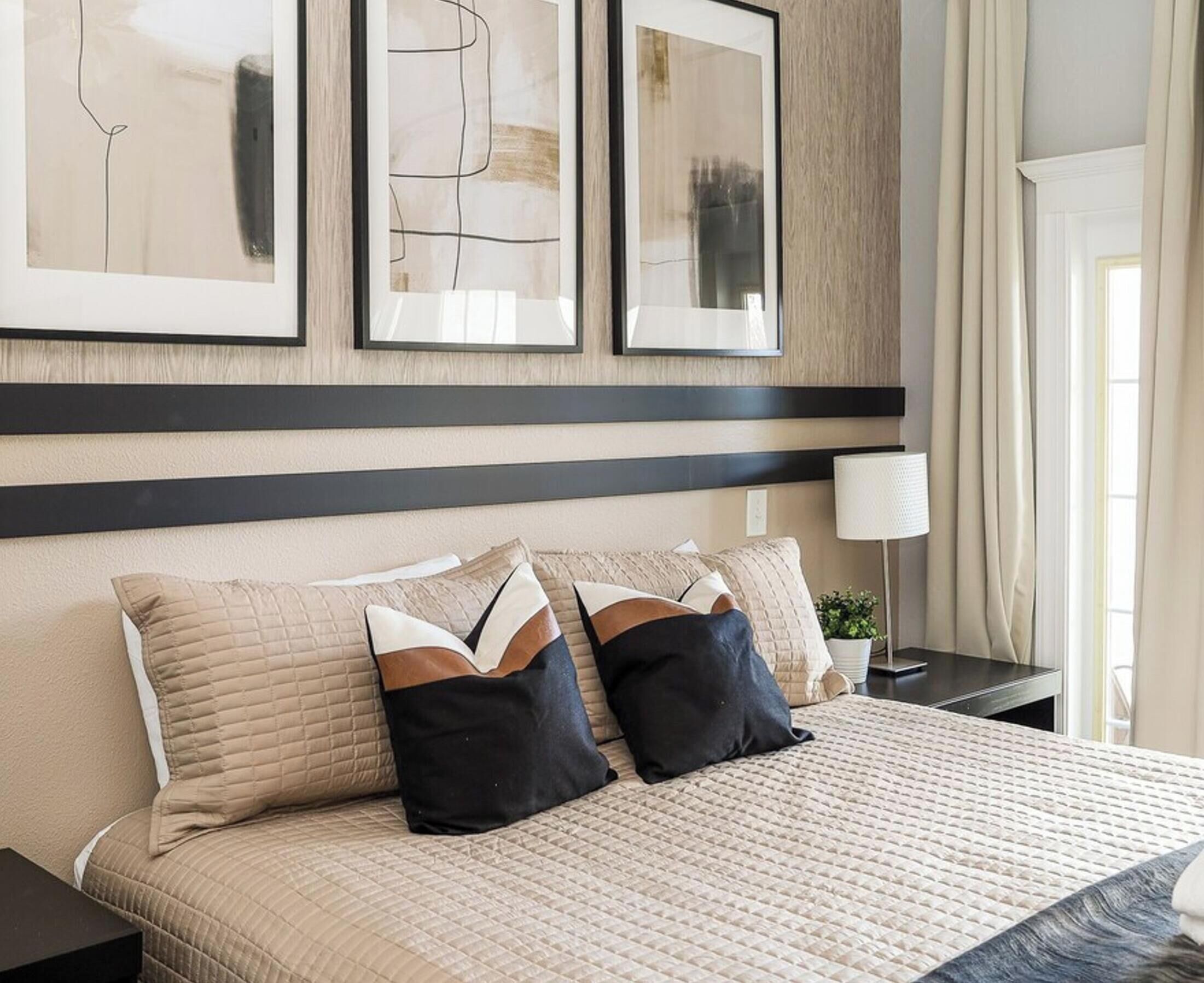 A hotel room with a bed, two bedside tables, a nightstand, a lamp, and three framed abstract artworks on the wall.