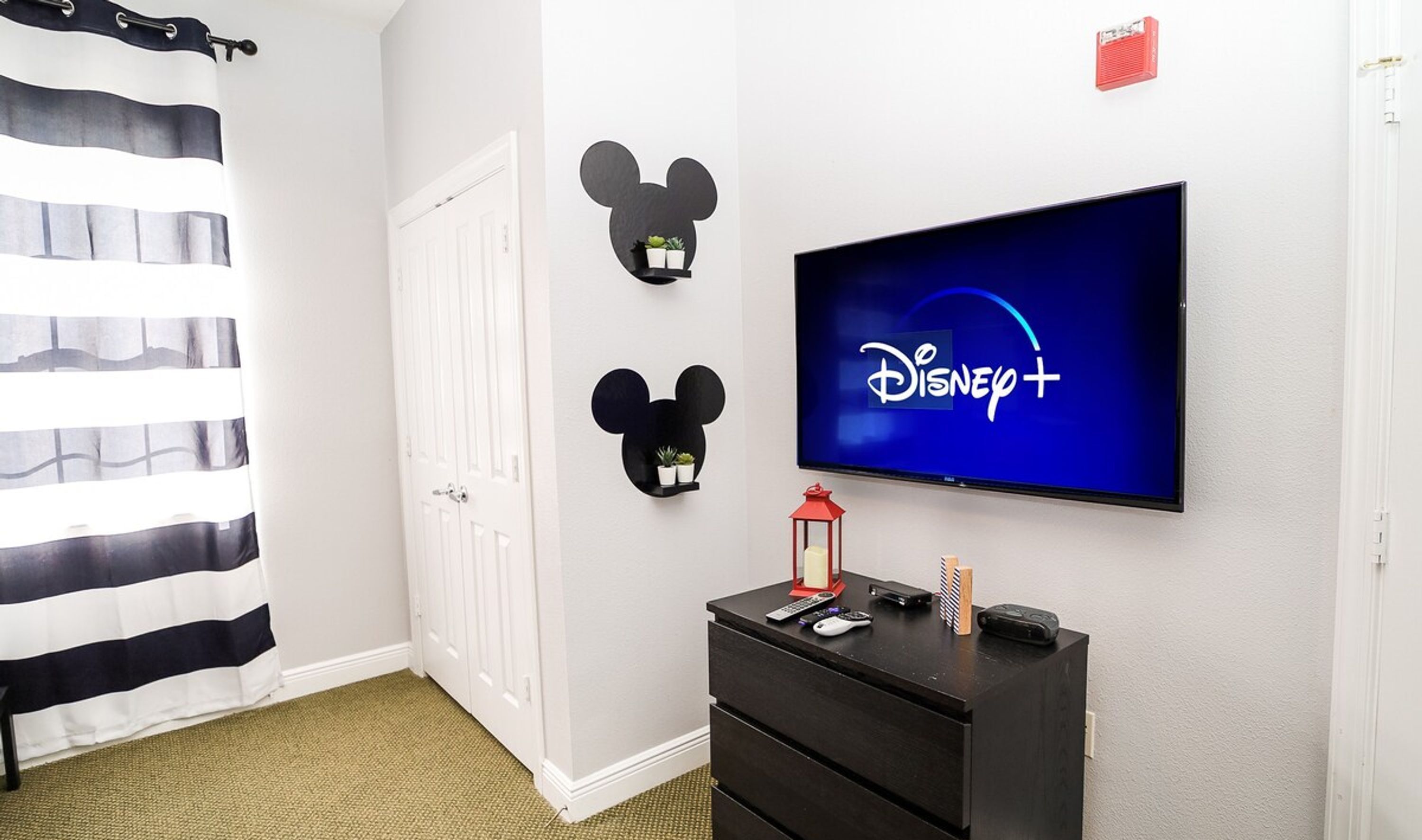 A hotel room with a television displaying the Disney+ logo, a black dresser, and two wall-mounted Mickey Mouse decorations.
