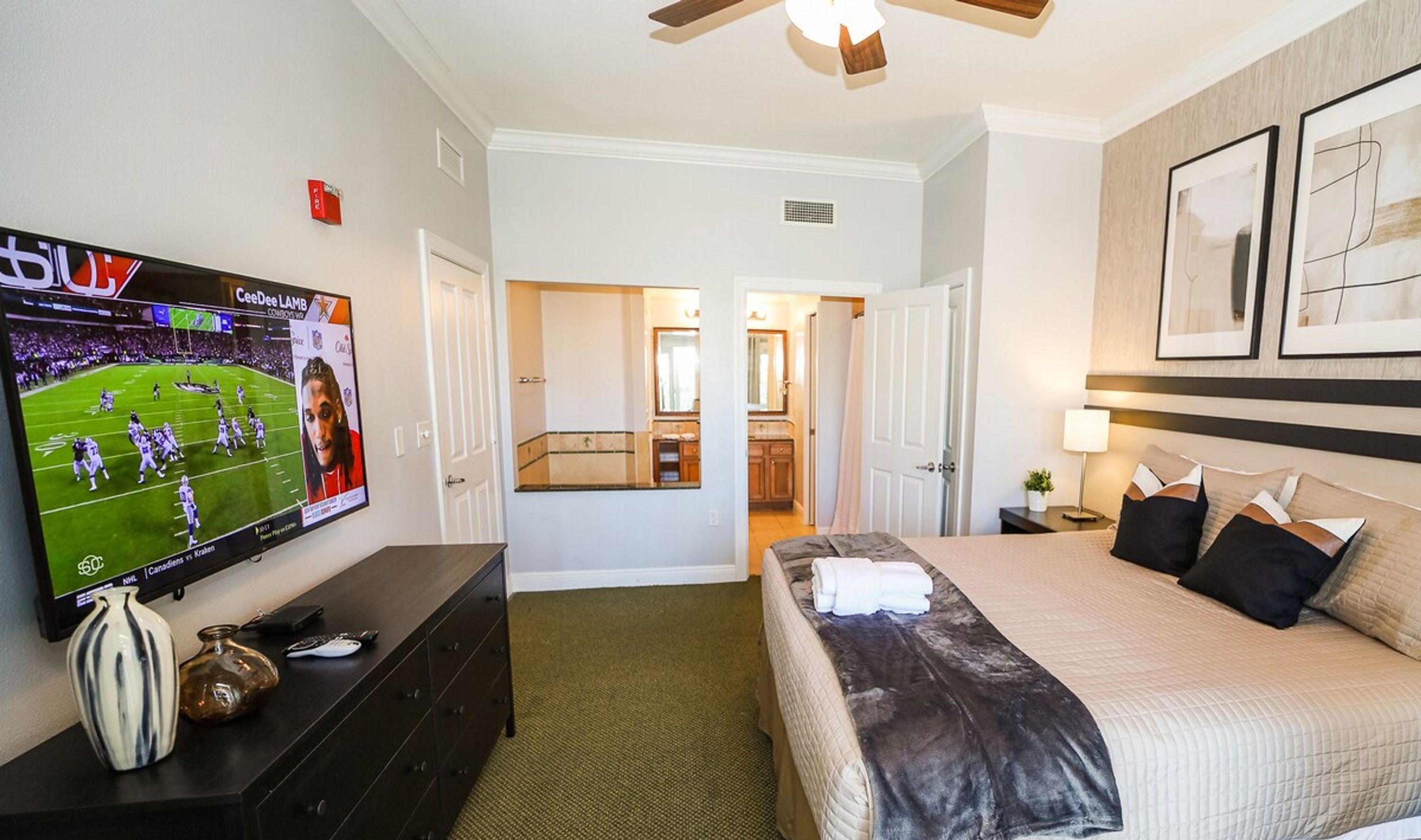 A hotel room with a bed, a TV displaying a sports game, a dresser, and a hallway leading to a bathroom.
