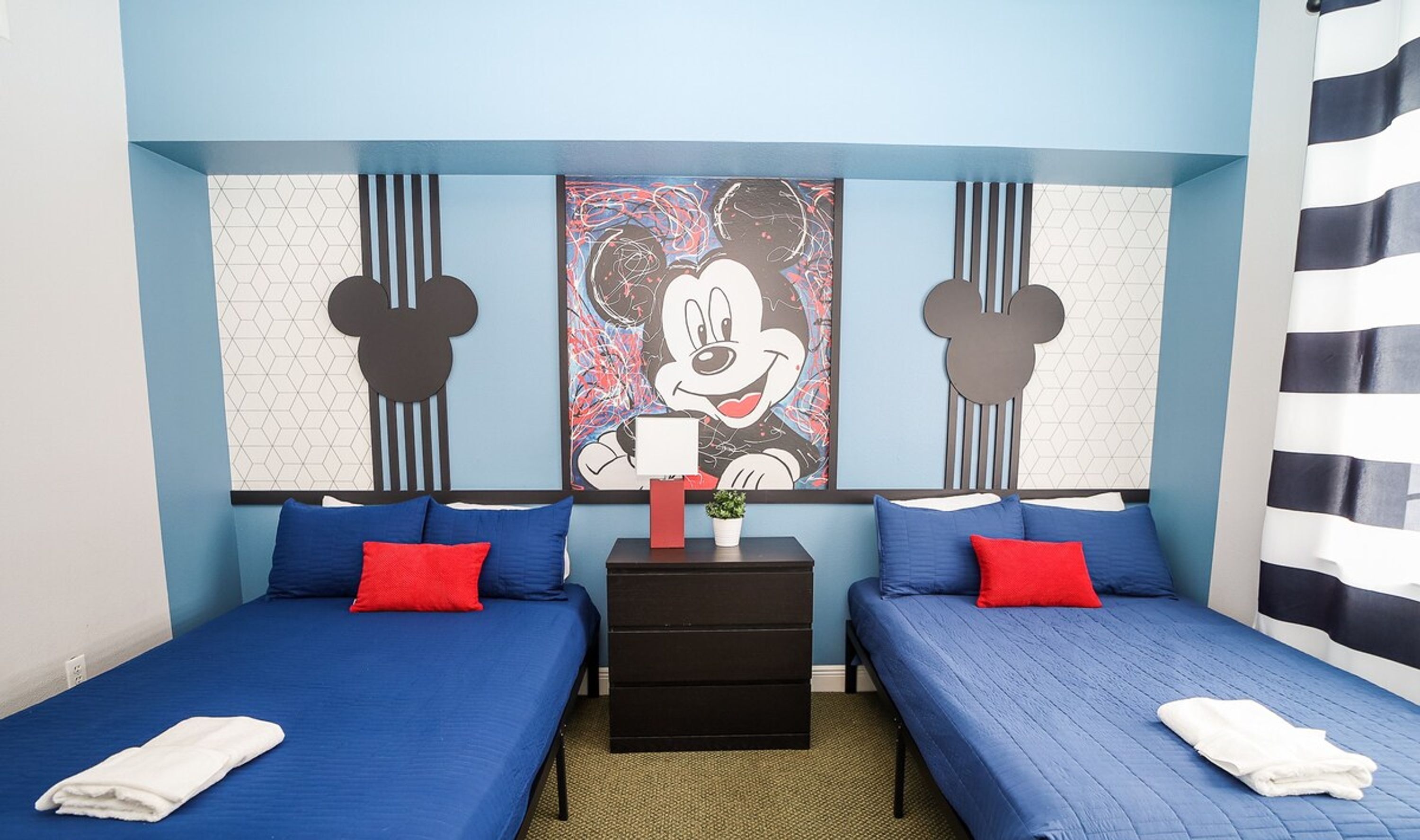 Habitación de hotel con dos camas, cada una con ropa de cama azul y almohadas rojas. En la pared, un cuadro de Mickey Mouse, una mesita con un objeto rojo y una mesita de noche negra.