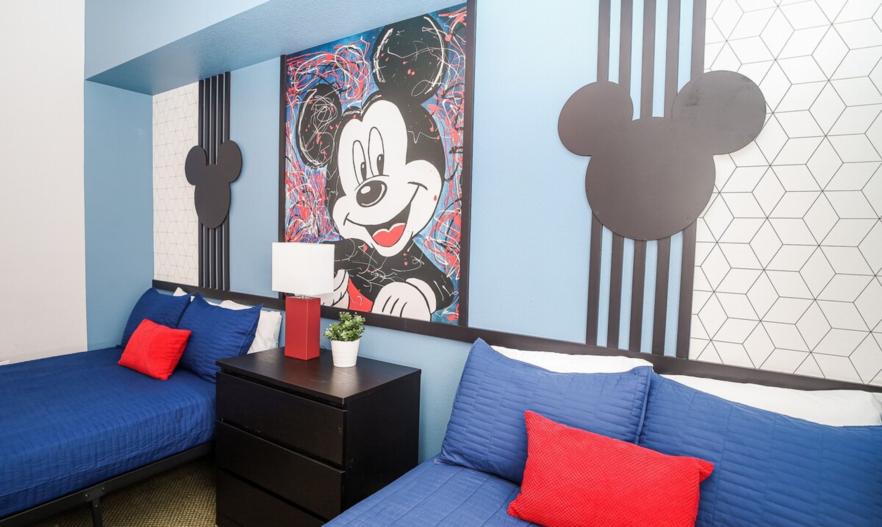 A hotel room with two beds, each with blue bedding and red pillows. There is a Mickey Mouse painting on the wall, a black nightstand with a lamp, and a geometric-patterned wall.
