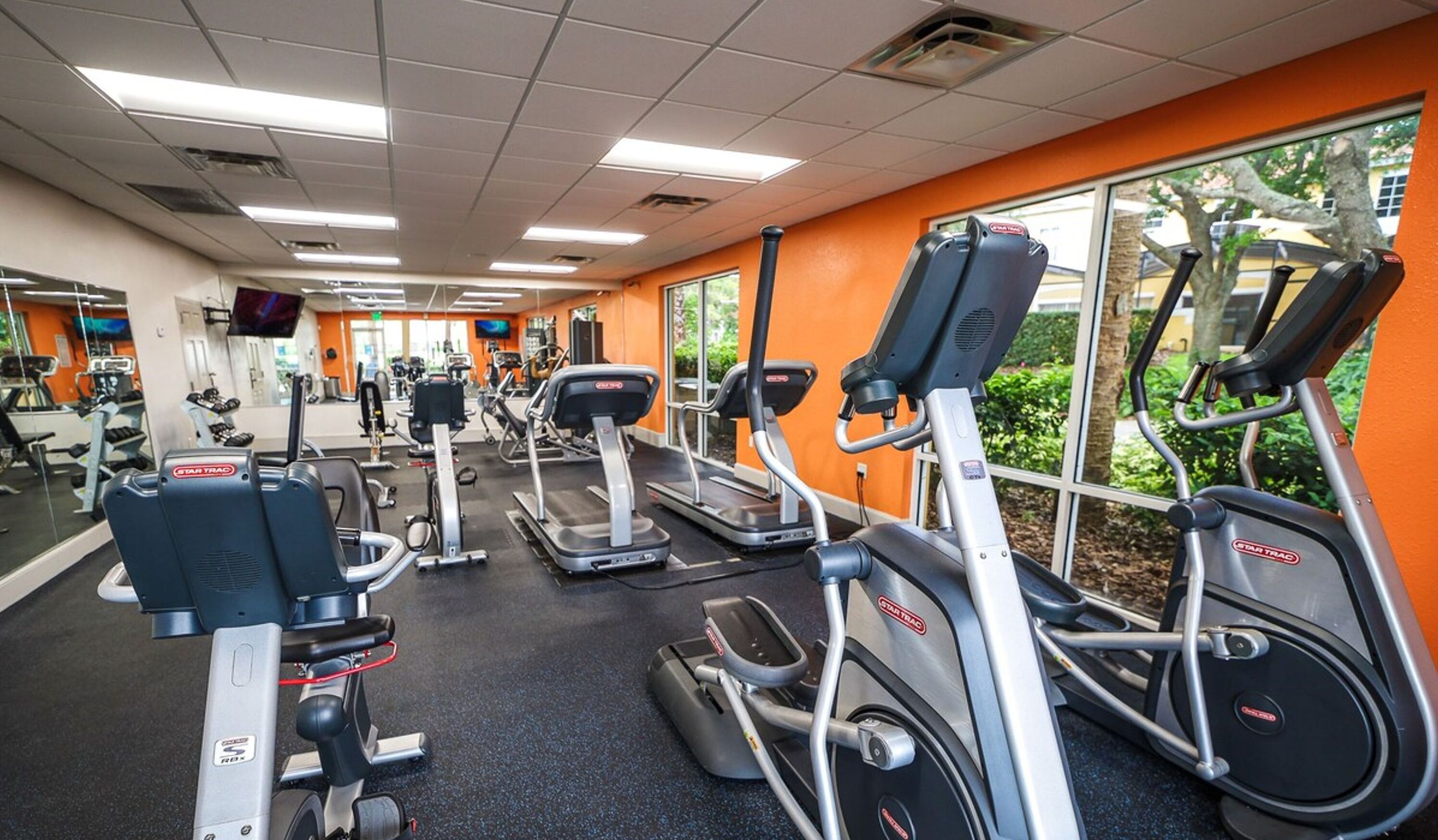 A gym with various cardio equipment, including treadmills and elliptical machines, and a large window providing a view of the outdoors.