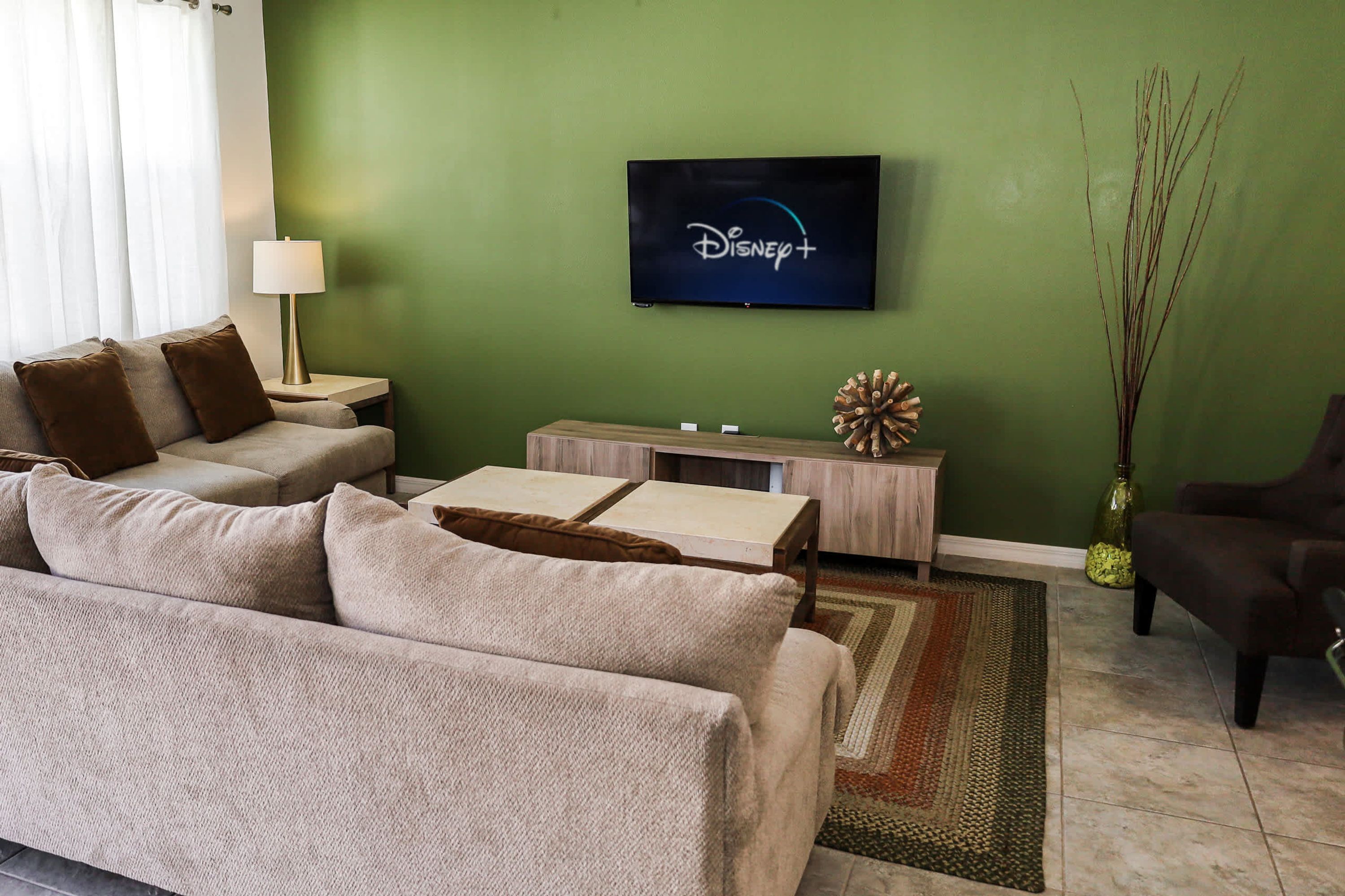 A living room with a green wall, a flat-screen TV displaying the Disney+ logo, a beige sofa, a small side table with a lamp, and a decorative vase.