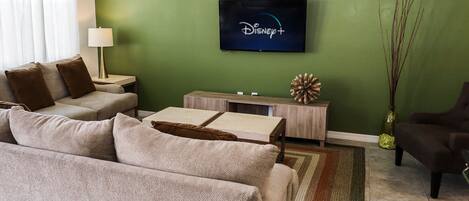 A living room with a green wall, a flat-screen TV displaying the Disney+ logo, a beige sofa, a small side table with a lamp, and a decorative vase.