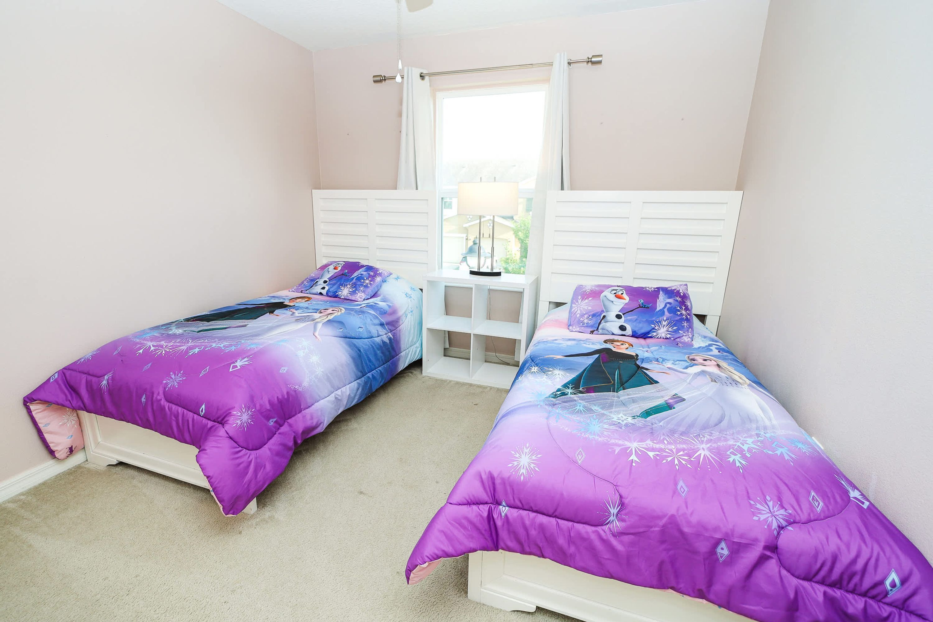A bedroom with two beds, each with a purple bedspread featuring a winter theme, white headboards, and a window with a white curtain.