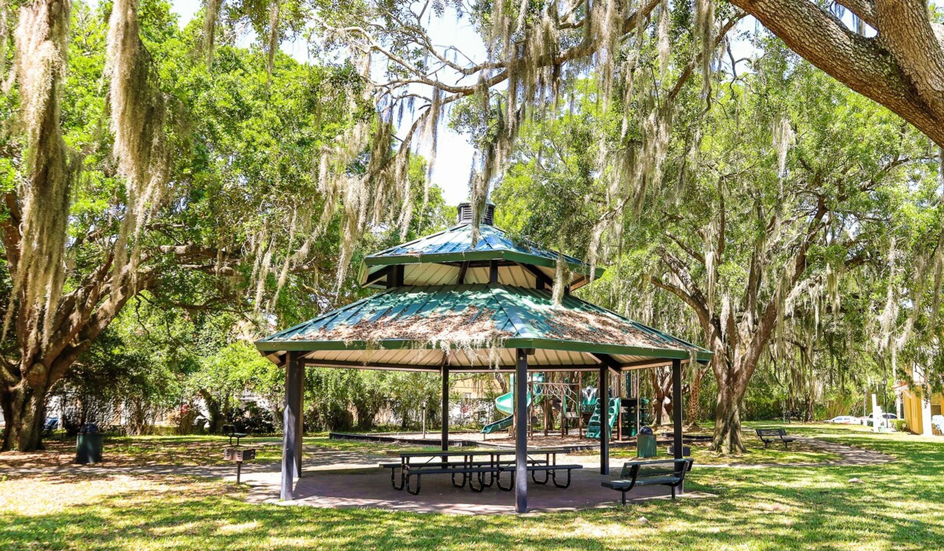 A gazebo with a green roof and metal frame, surrounded by trees with Spanish moss, benches, and picnic tables in a park.