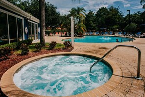A hot tub with a tiled edge, surrounded by a brick patio, and a pool area with lounge chairs and palm trees.