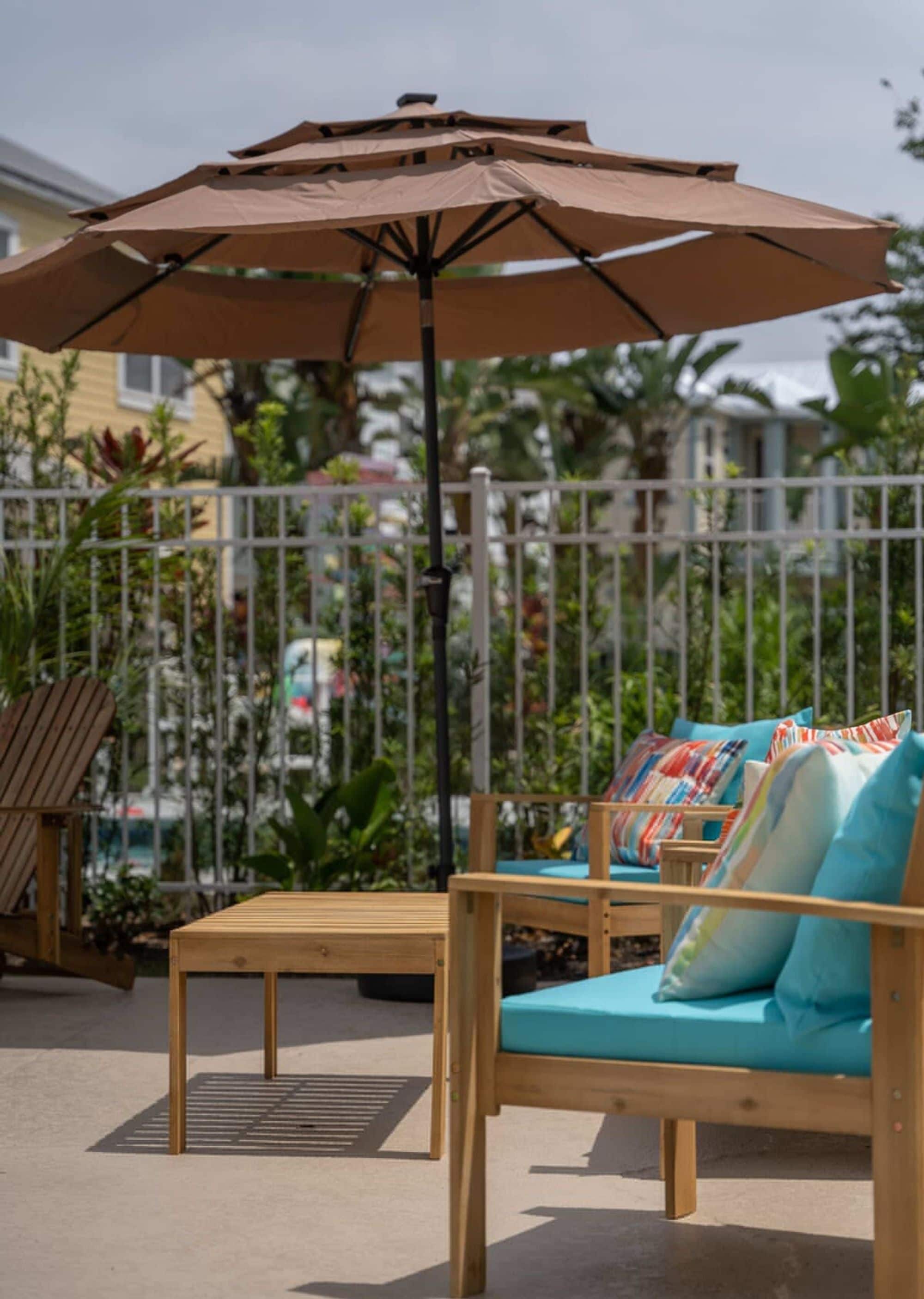 A wooden outdoor seating area with a large umbrella, a metal fence, and tropical plants in the background.