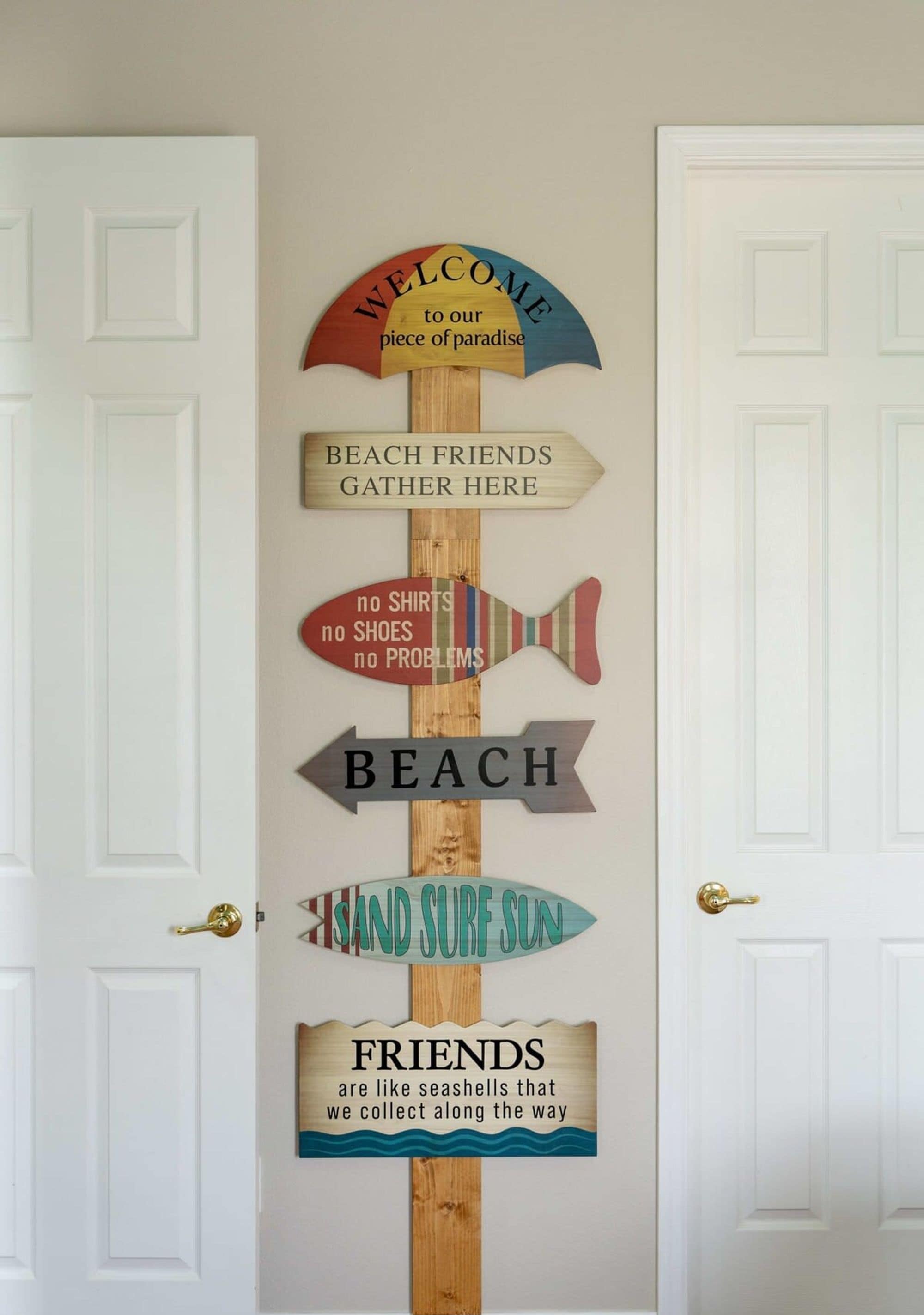 Un letrero de madera con varios carteles y mensajes, incluyendo 'BIENVENIDOS a nuestro pedazo de paraíso' y 'AMIGOS DE LA PLAYA SE REÚNEN AQUÍ'.