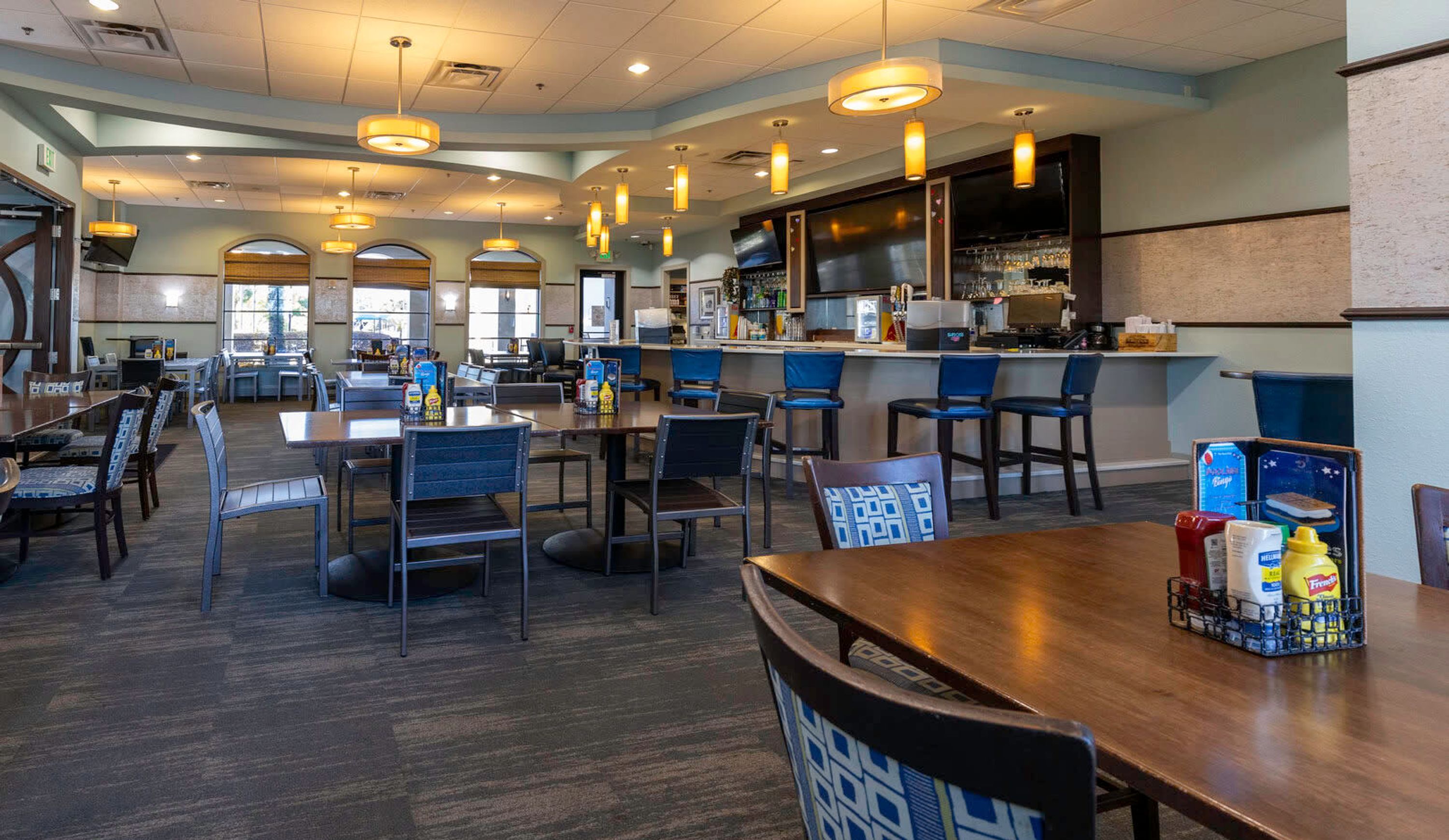 A spacious restaurant with multiple dining tables and chairs, a bar area, and a kitchen visible in the background.