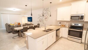 Una cocina moderna con armarios blancos, una isla central, electrodomésticos de acero inoxidable y un área de comedor con un sofá y sillas grises.