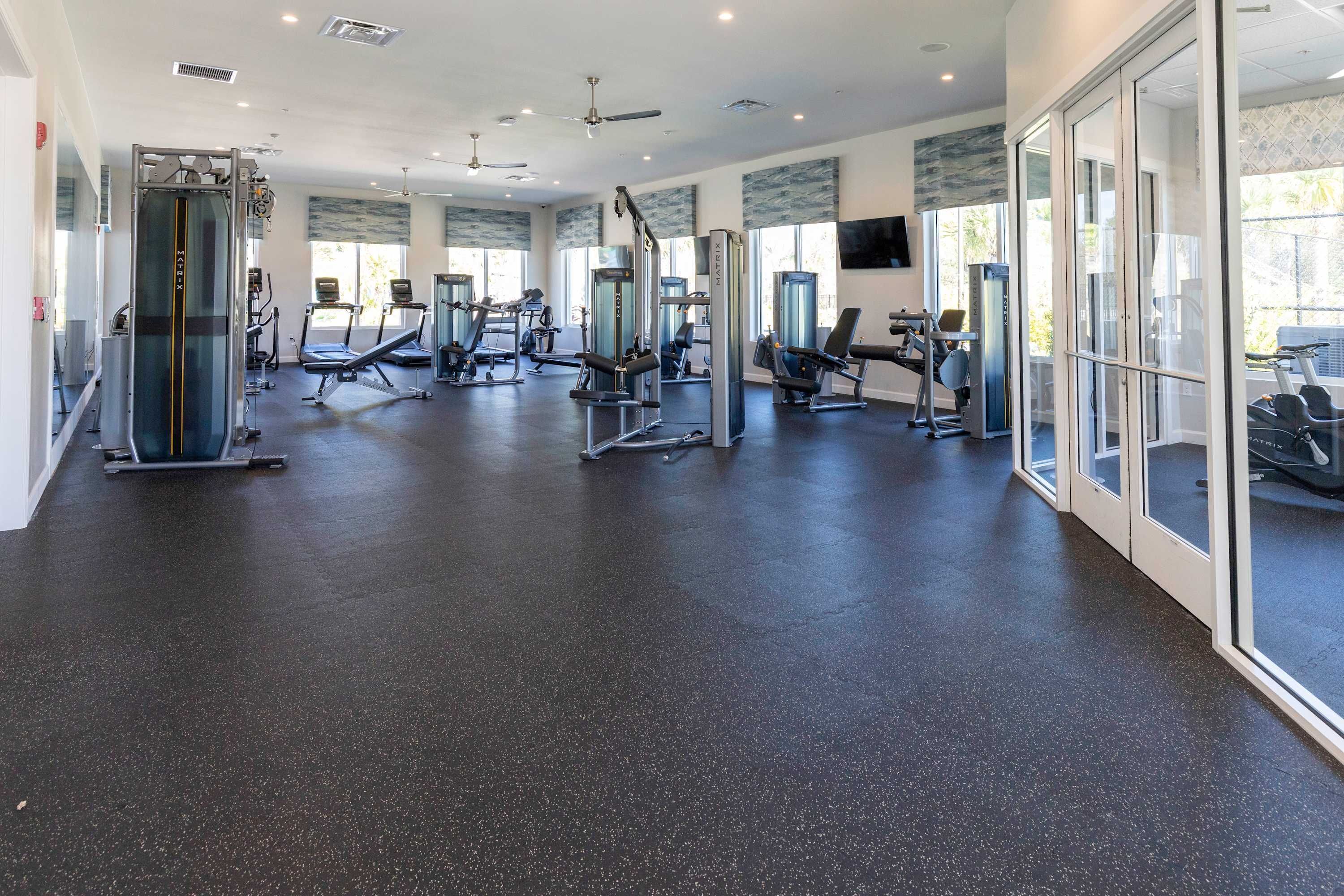 Ein Fitnessstudio mit verschiedenen Trainingsgeräten, großen Fenstern und einem Deckenventilator.
