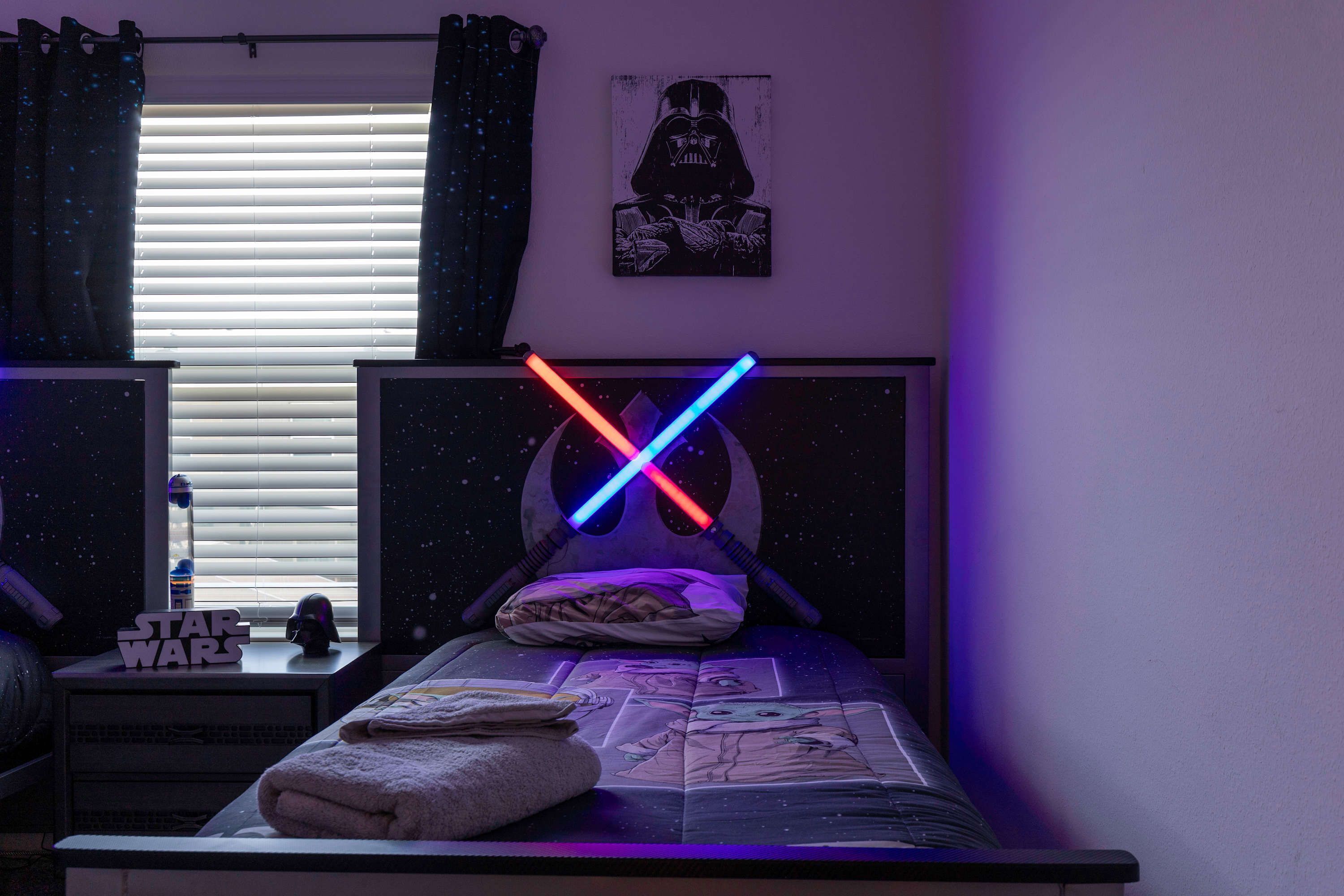 A bedroom with a Star Wars themed bedspread, a Darth Vader poster on the wall, and a Star Wars sign on the nightstand.